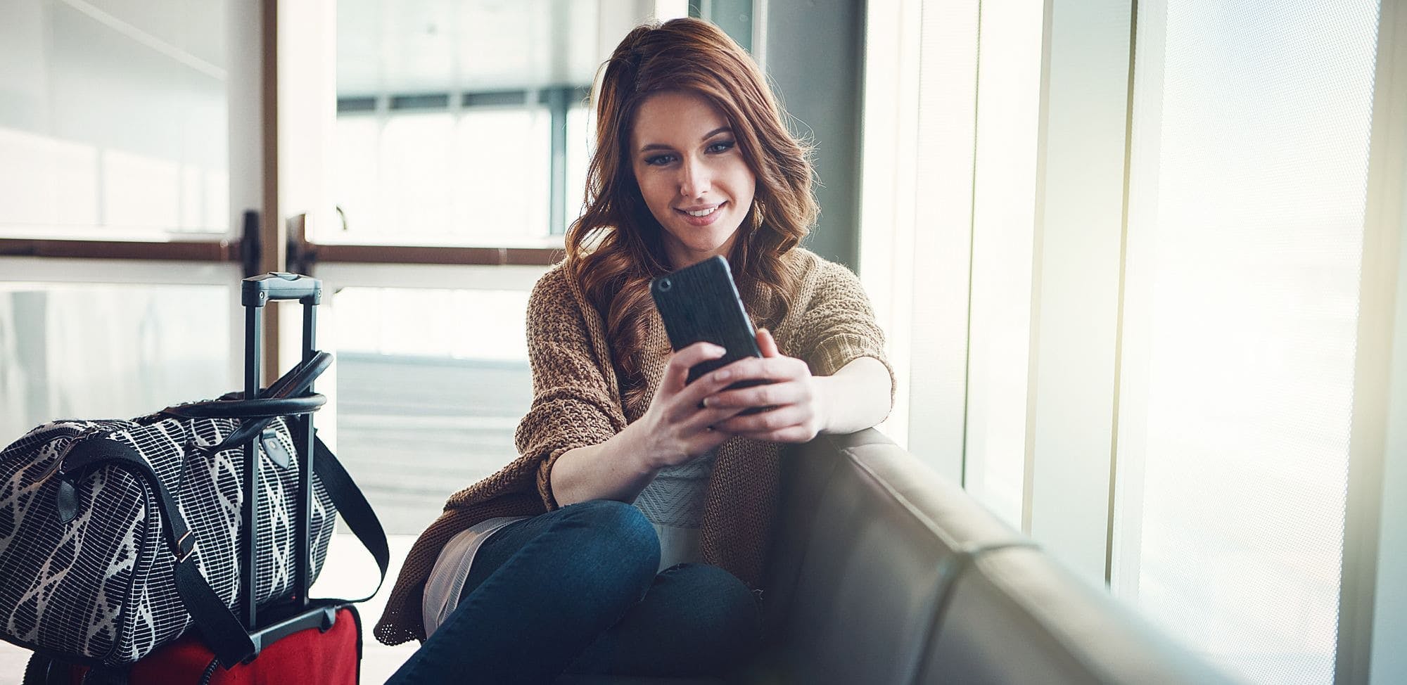 Une femme assise dans un aéroport, souriant tout en regardant son téléphone. À côté d'elle, une valise rouge avec un sac à main noir et blanc posé dessus. La lumière naturelle filtre à travers les fenêtres derrière elle.