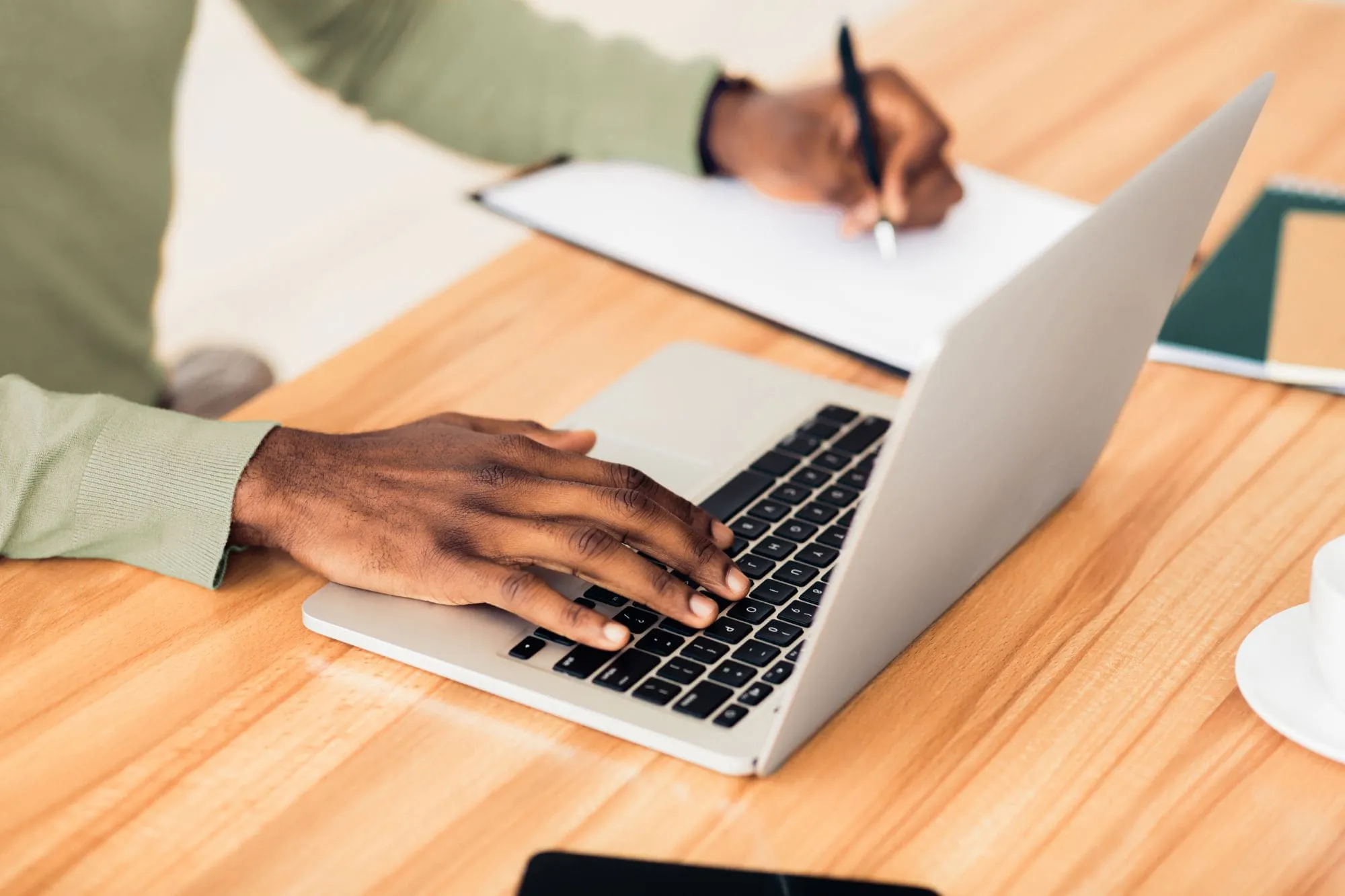 An unrecognizable Afro man working on his laptop while taking notes on a notebook beside him. A coffee cup is visible in the bottom right corner.