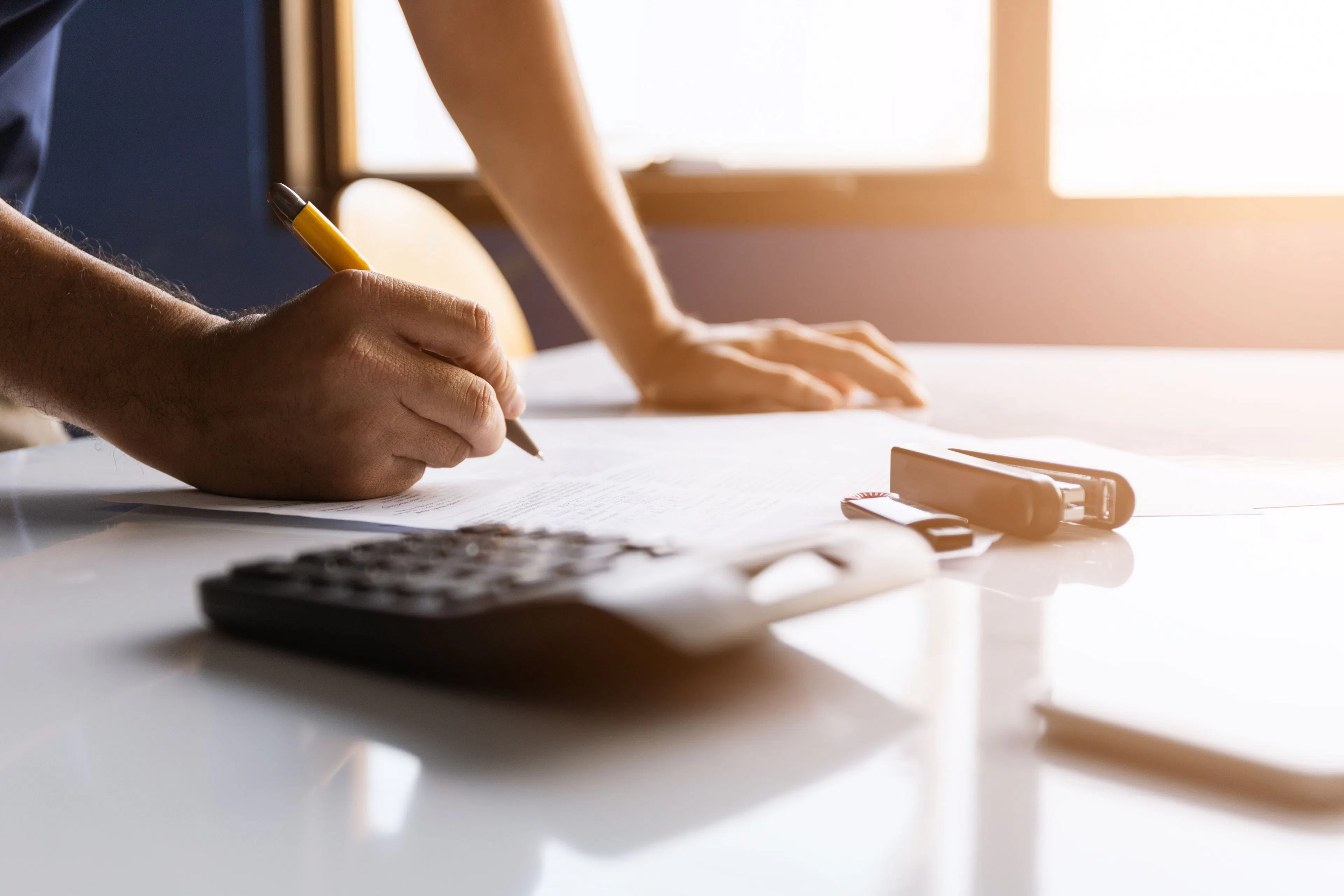 A businessman working in an office, taking notes on a document placed on a sunlit desk, with a calculator and a stapler in the foreground.