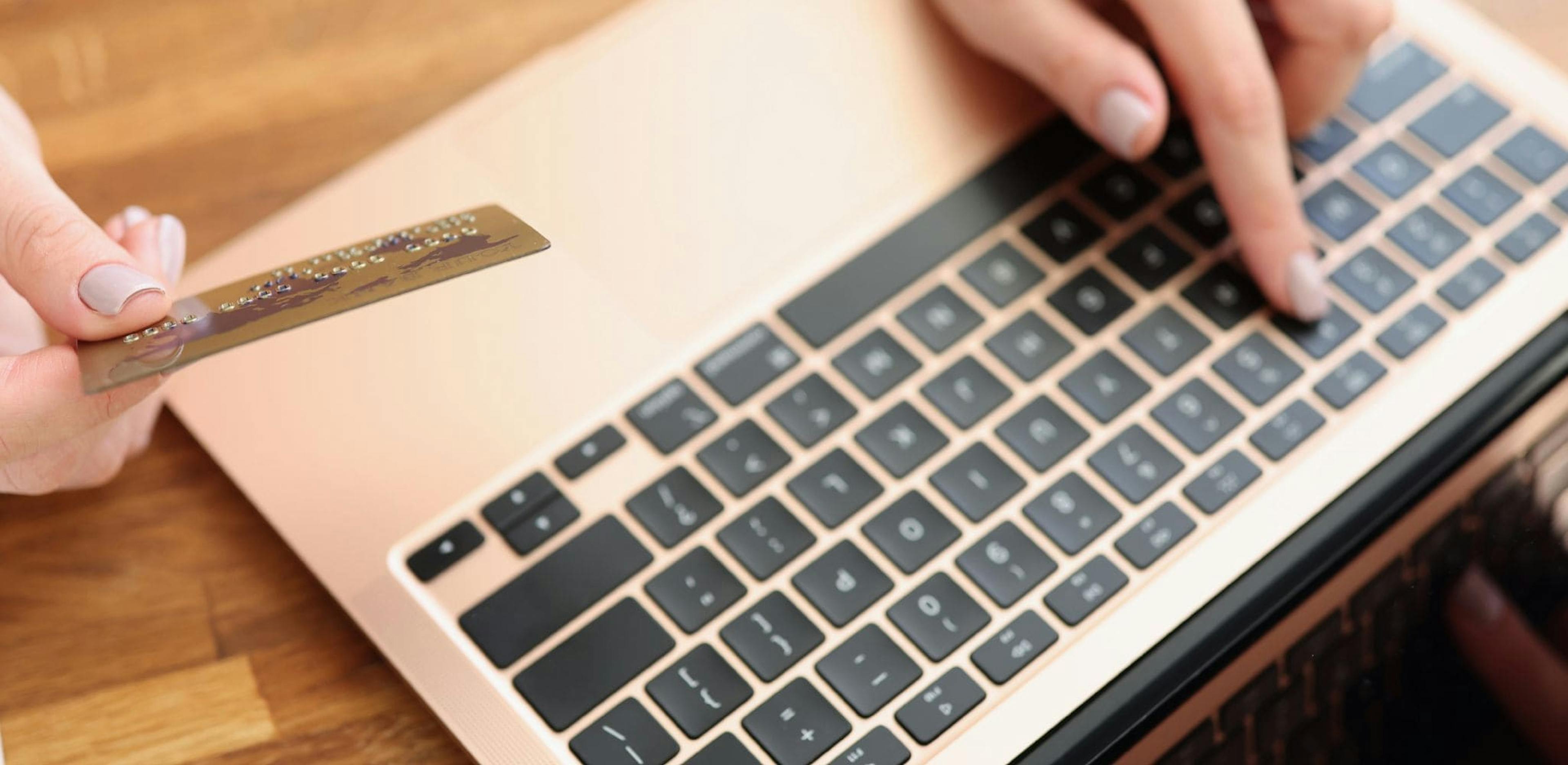 Close-up view of a person's hands typing on a gold laptop keyboard while holding a credit card. The image suggests an online payment or digital transaction in progress.