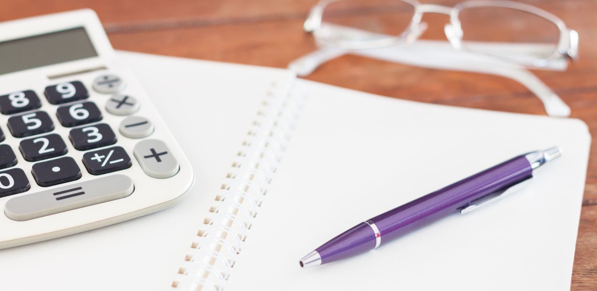 Vue de dessus d'un carnet à spirale ouvert, accompagné d'un stylo violet, d'une calculatrice et d'une paire de lunettes posée sur une table en bois. L'image suggère un espace de travail ou d'étude.