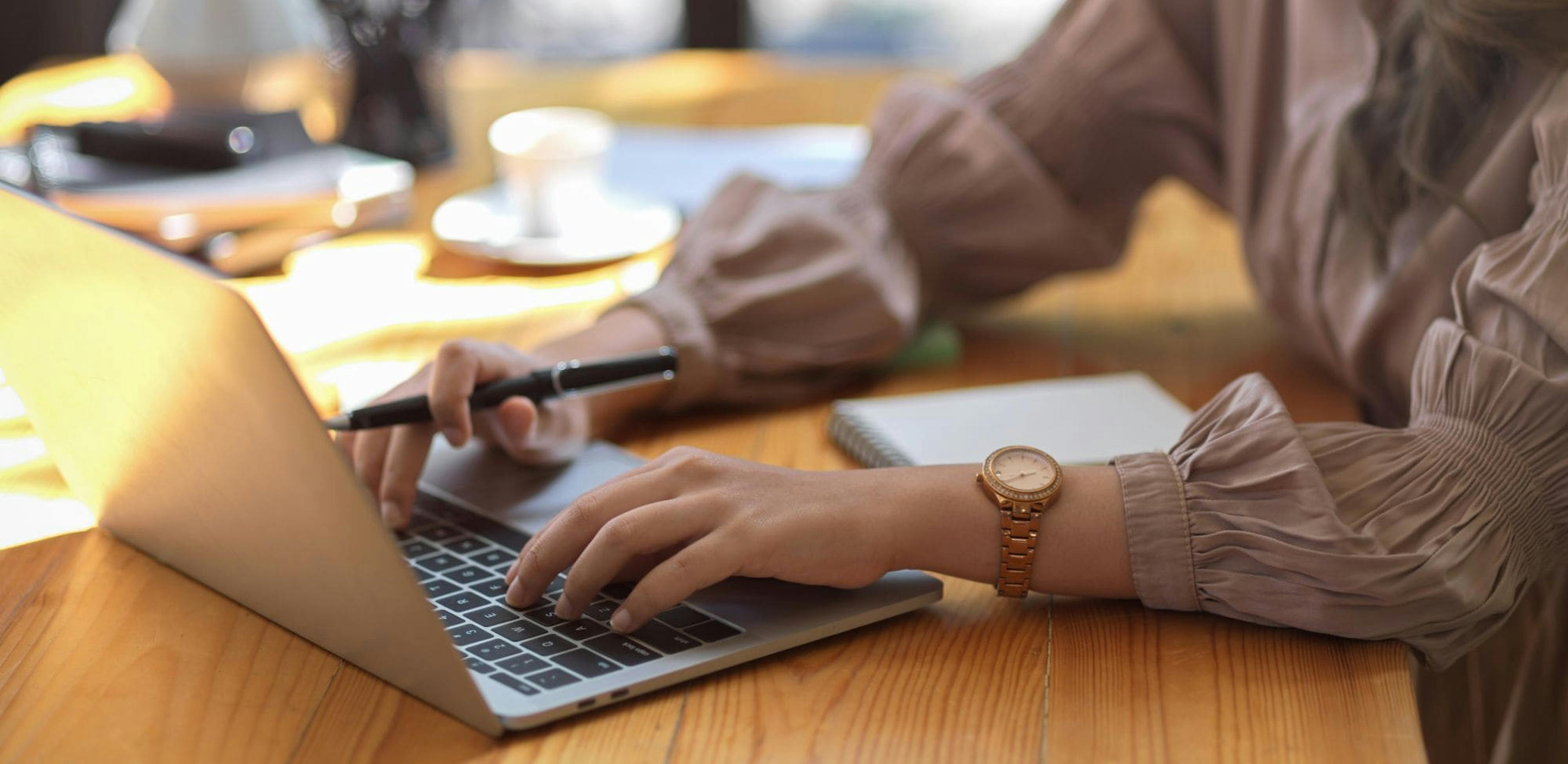 Partial view of a woman using a laptop on a wooden table. She is holding a pen in one hand while typing on the keyboard with the other. An open notebook and a cup of coffee are visible in the background.