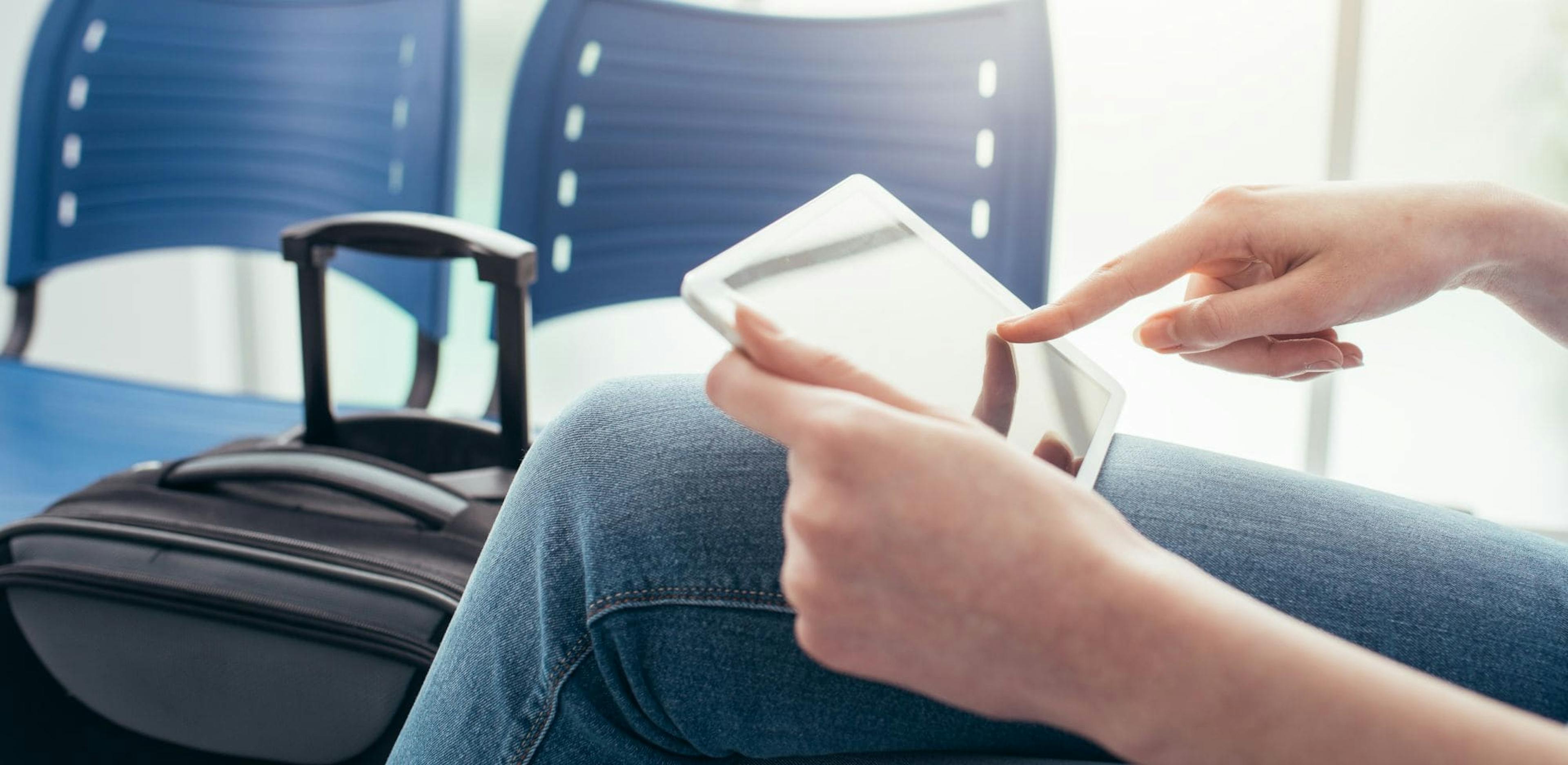 A person sitting in a waiting area, using a tablet. Next to them is a black suitcase. The seats around them are blue, suggesting they are in an airport terminal.