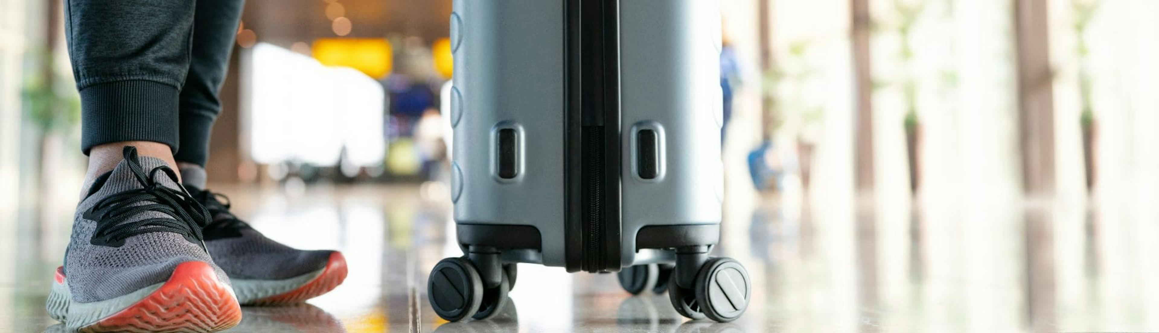 Close-up view of a person’s legs in casual attire, pulling a wheeled suitcase in an airport terminal. The shiny floor and soft lighting suggest a modern travel environment.