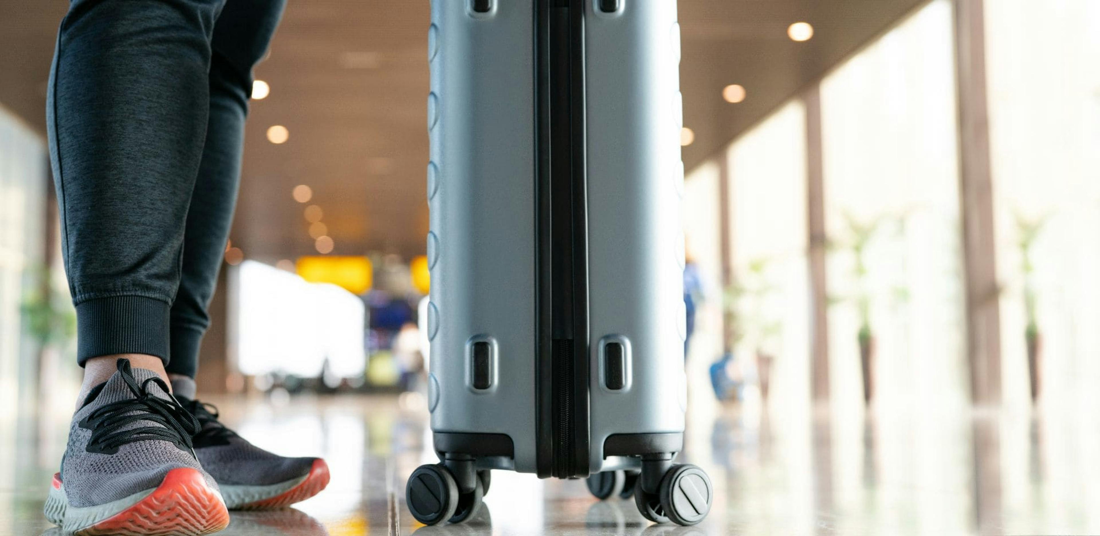 Close-up view of a person’s legs in casual attire, pulling a wheeled suitcase in an airport terminal. The shiny floor and soft lighting suggest a modern travel environment."