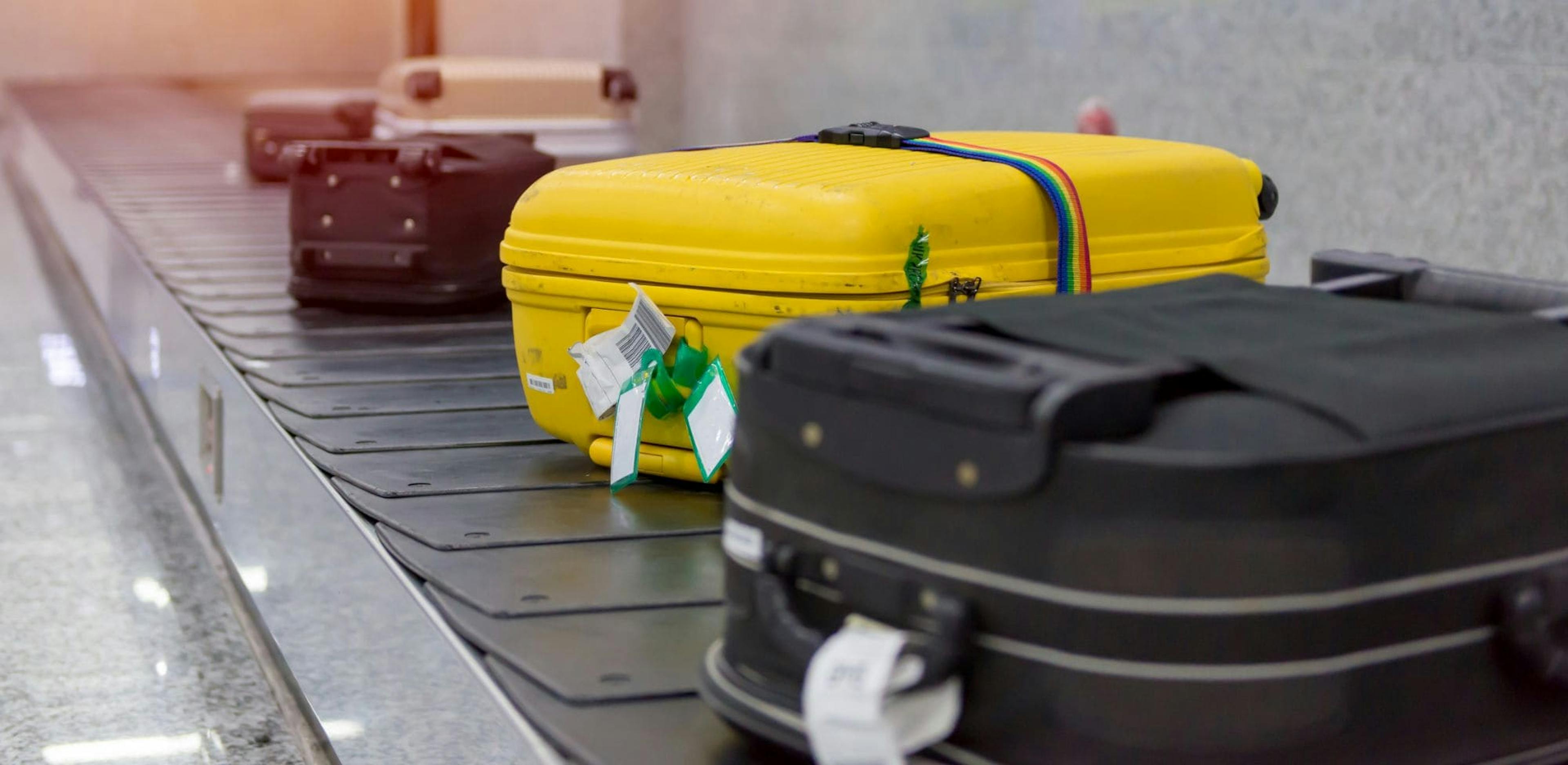 Close-up view of several suitcases on an airport baggage carousel. Among the luggage, a bright yellow suitcase with a rainbow strap stands out among the darker bags.