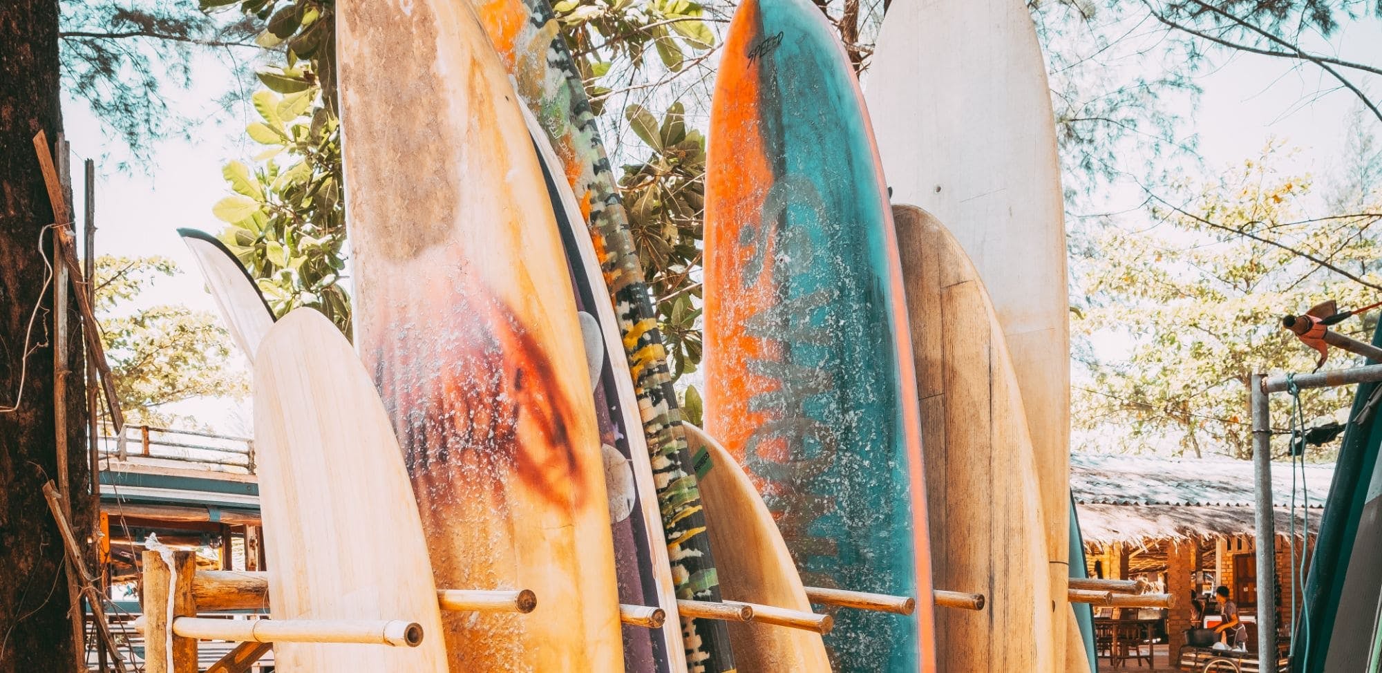 Un ensemble de planches de surf colorées empilées les unes contre les autres, disponibles à la location sur une plage. Les planches, de différentes couleurs et tailles, sont rangées à l'extérieur sous les arbres, créant une atmosphère tropicale.