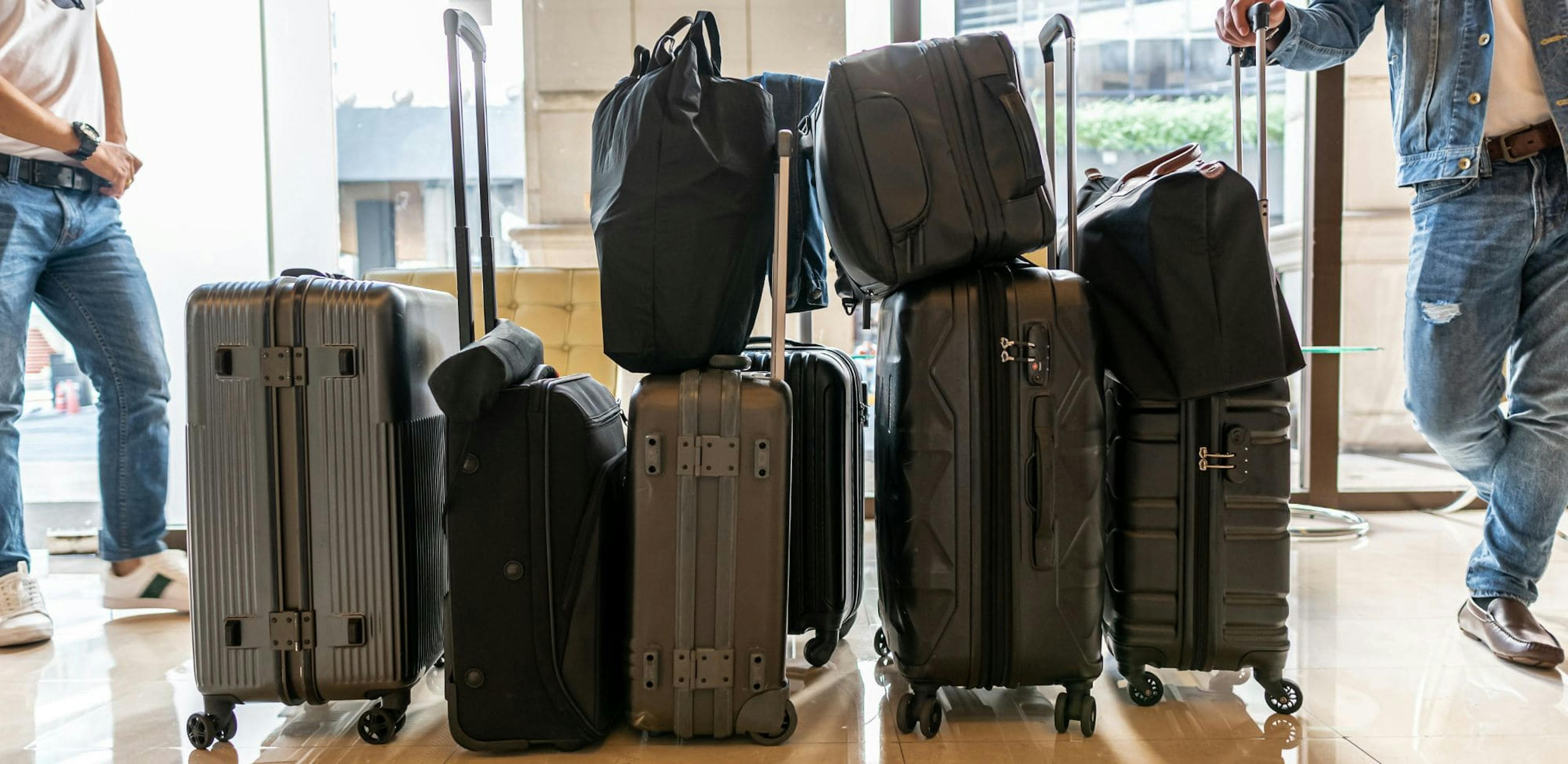 Partial view of several men standing with suitcases at an airport. The luggage is lined up and ready for travel. The men are wearing jeans and casual shoes.