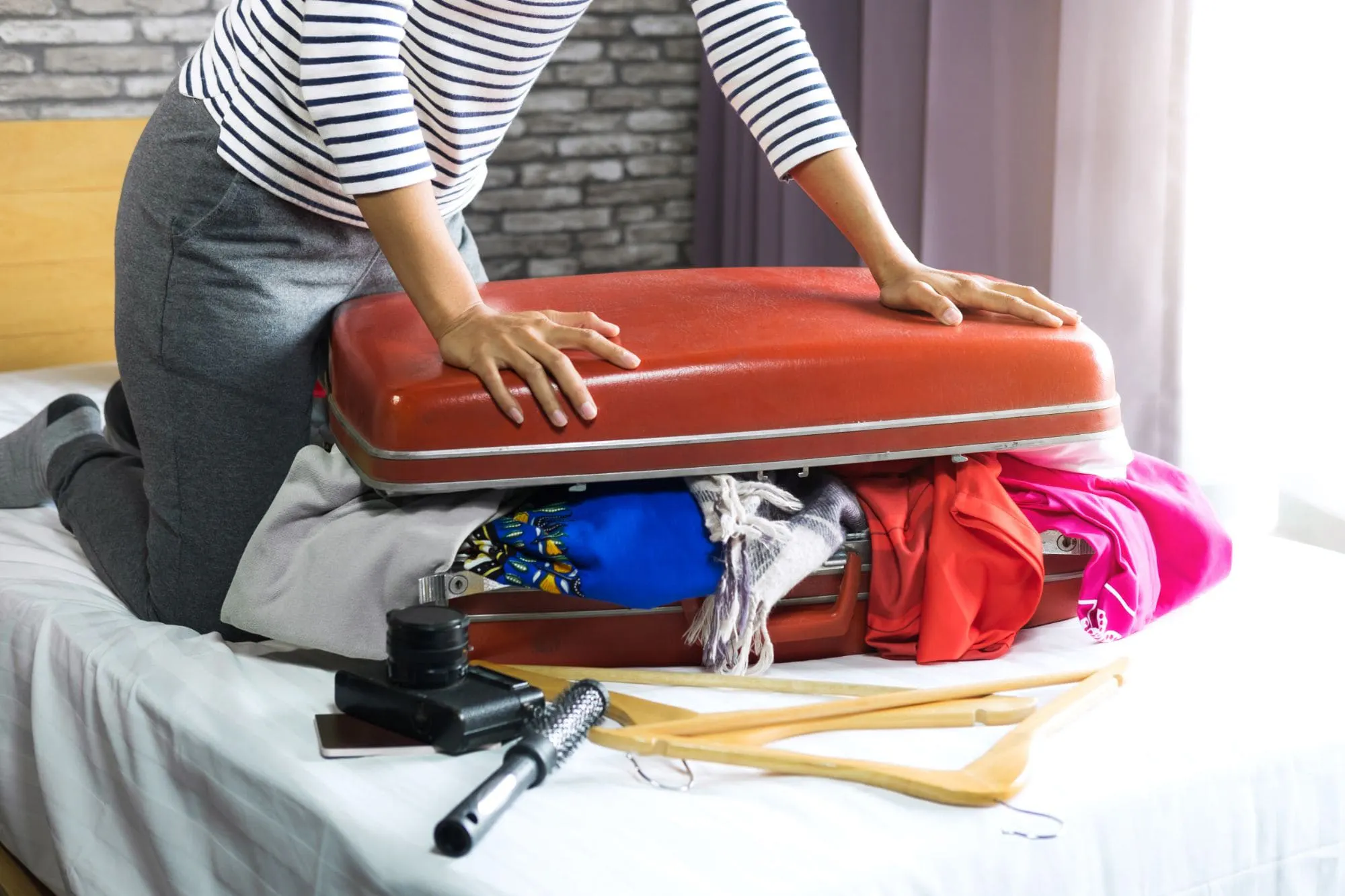 Low angle view of a woman trying to close an overstuffed suitcase placed on a bed, with clothes spilling out the sides and various travel items scattered nearby.