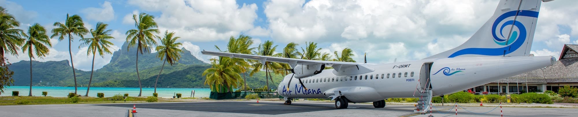 Avion de la compagnie Air Moana stationné sur le tarmac d'un aéroport tropical, entouré de palmiers avec une vue sur l'océan turquoise et des montagnes en arrière-plan sous un ciel nuageux.