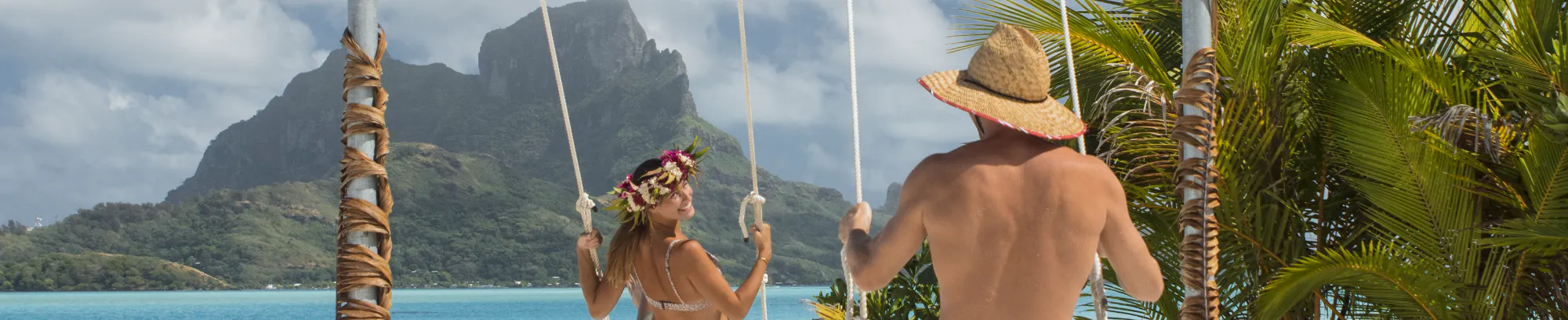 Un homme et une femme, tous deux en maillot de bain, se balancent sur des balançoires face à la mer turquoise. La femme porte une couronne de fleurs et l'homme un chapeau de paille. En arrière-plan, des montagnes verdoyantes et un ciel partiellement nuageux.