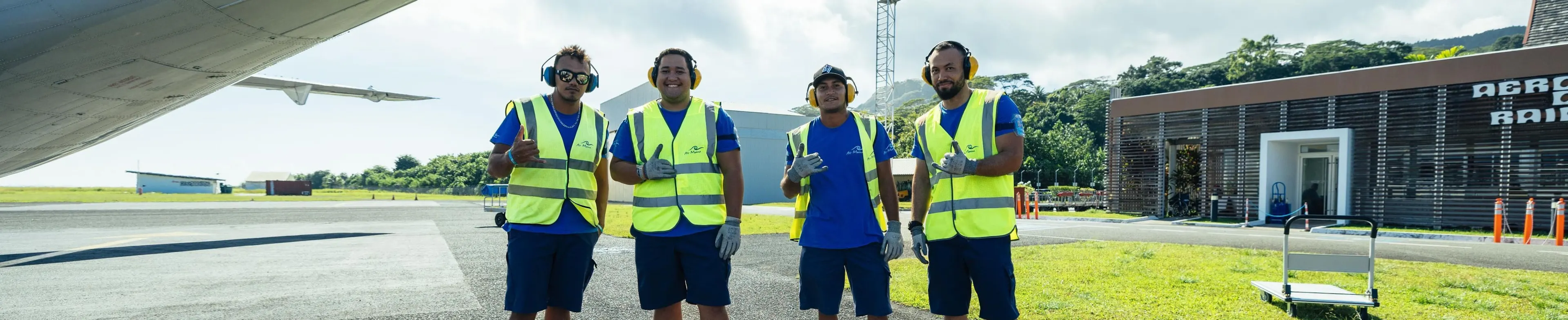 Quatre travailleurs portant des gilets de sécurité jaunes et des protections auditives posent en souriant sur le tarmac d'un aéroport.
