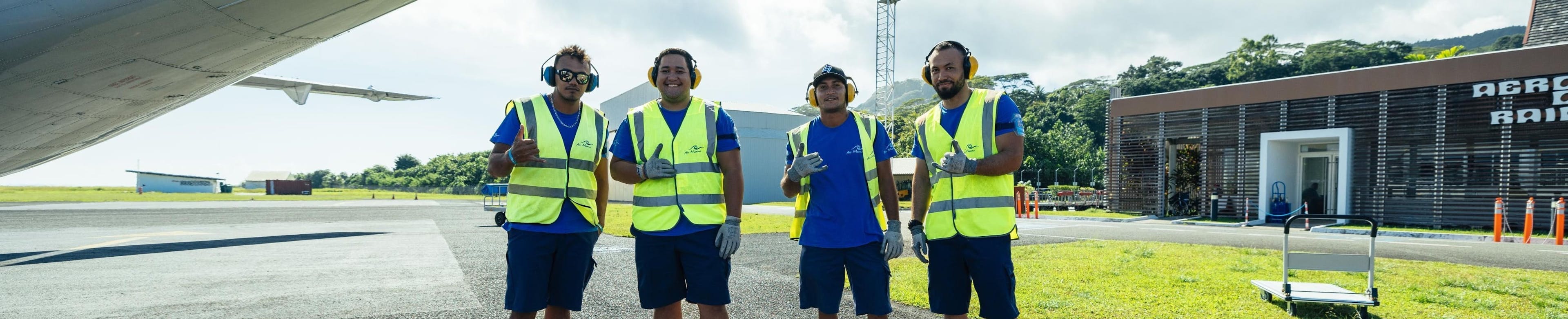 Four workers wearing yellow safety vests and ear protection pose smiling on an airport tarmac.