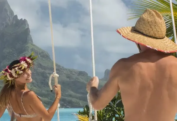 A man and a woman, both in swimsuits, are swinging on swings facing the turquoise sea. The woman is wearing a flower crown and the man a straw hat. In the background, lush green mountains and a partly cloudy sky.