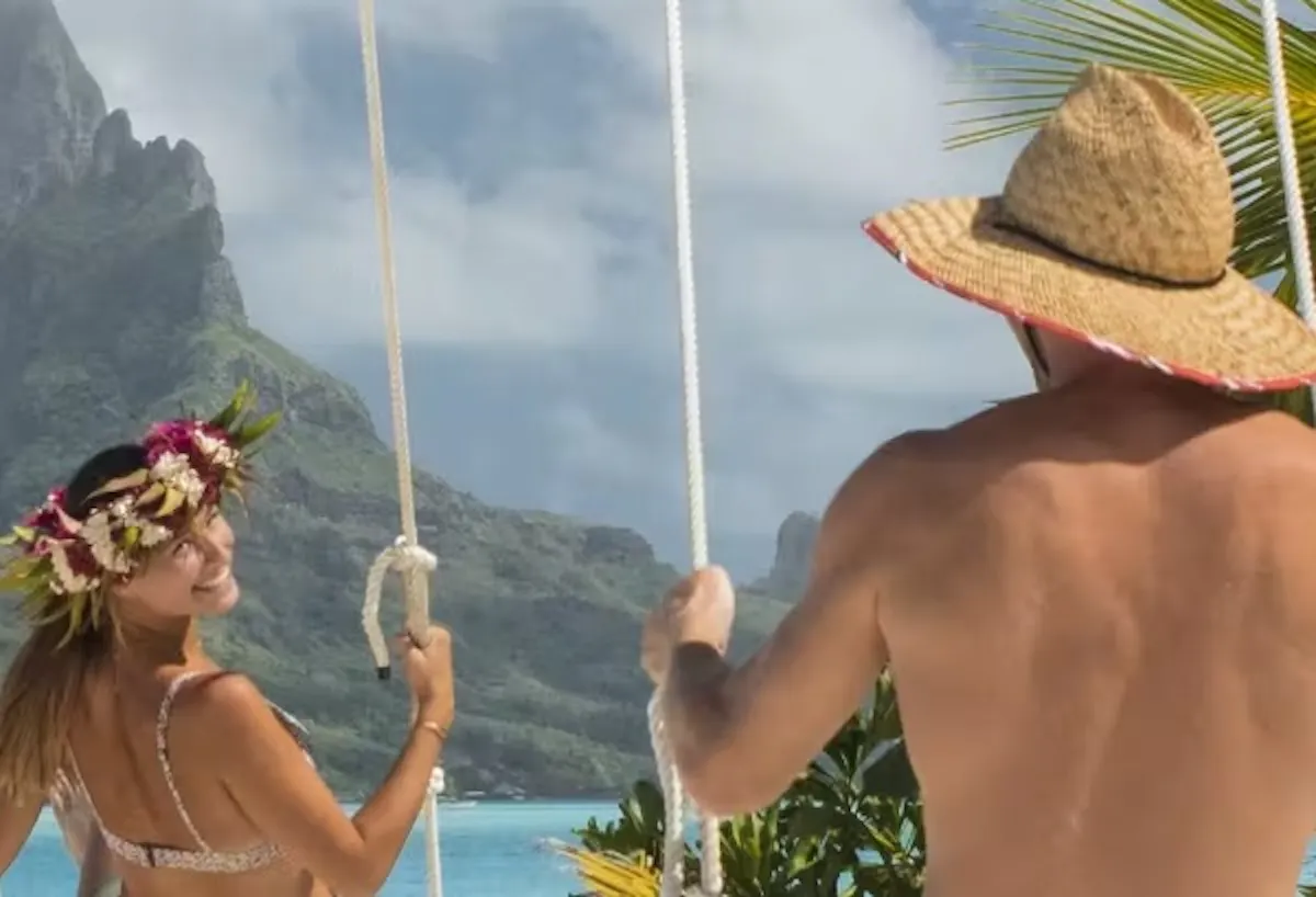 A man and a woman, both in swimsuits, are swinging on swings facing the turquoise sea. The woman is wearing a flower crown and the man a straw hat. In the background, lush green mountains and a partly cloudy sky.