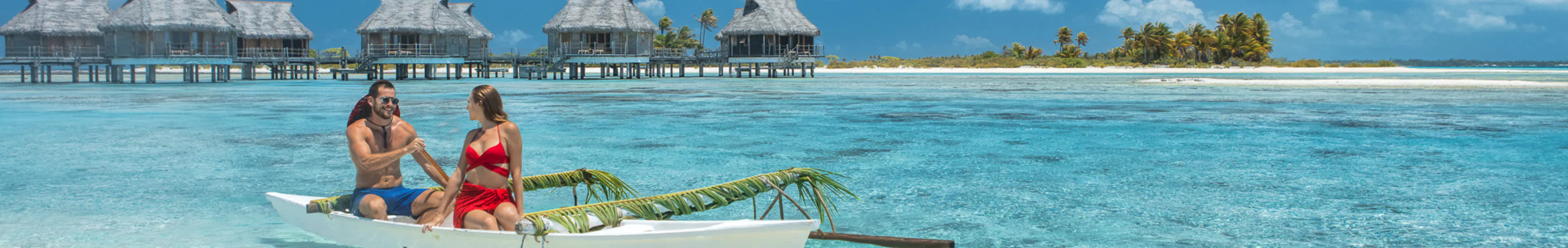 Un couple assis dans une pirogue traditionnelle sur un lagon turquoise en Polynésie française, avec des bungalows sur pilotis et une petite île bordée de palmiers à l’arrière-plan sous un ciel bleu.