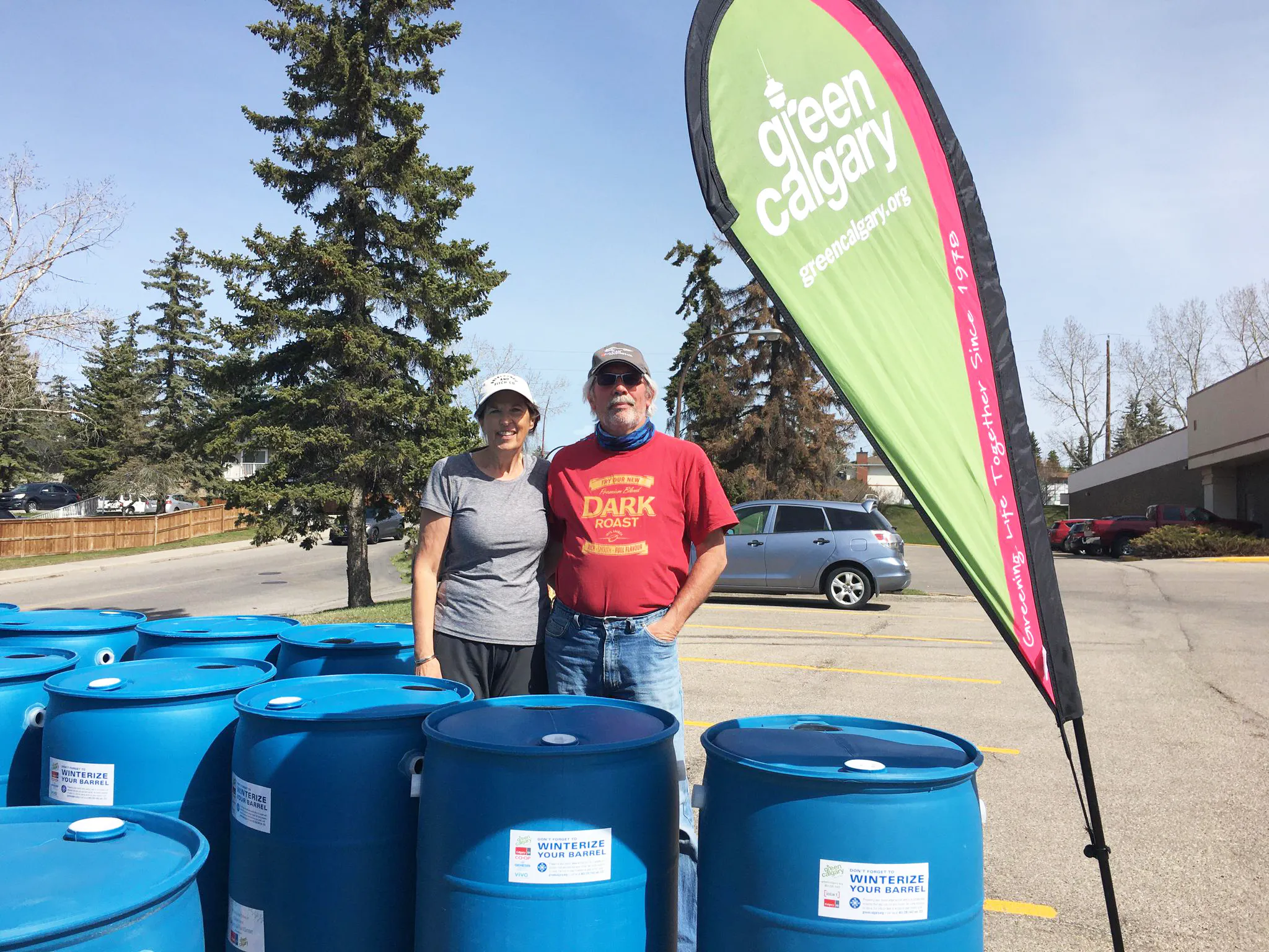 Two people standing beside a Green Calgary flag and rain barrels.