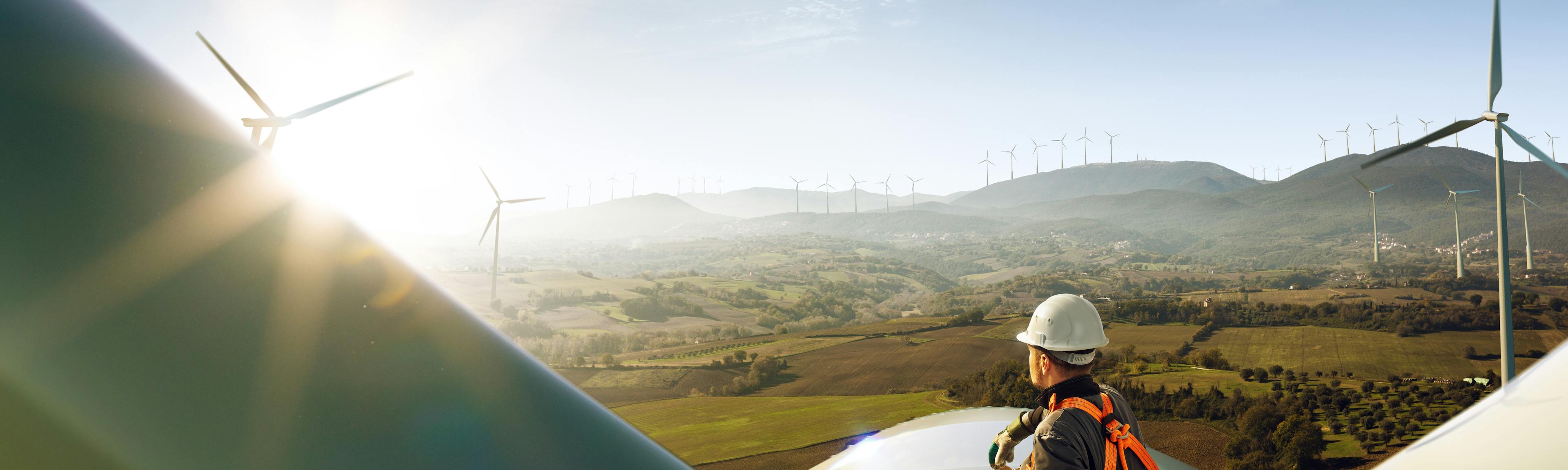 A man in a hard hat sits on top of a windmill in a windfarm.