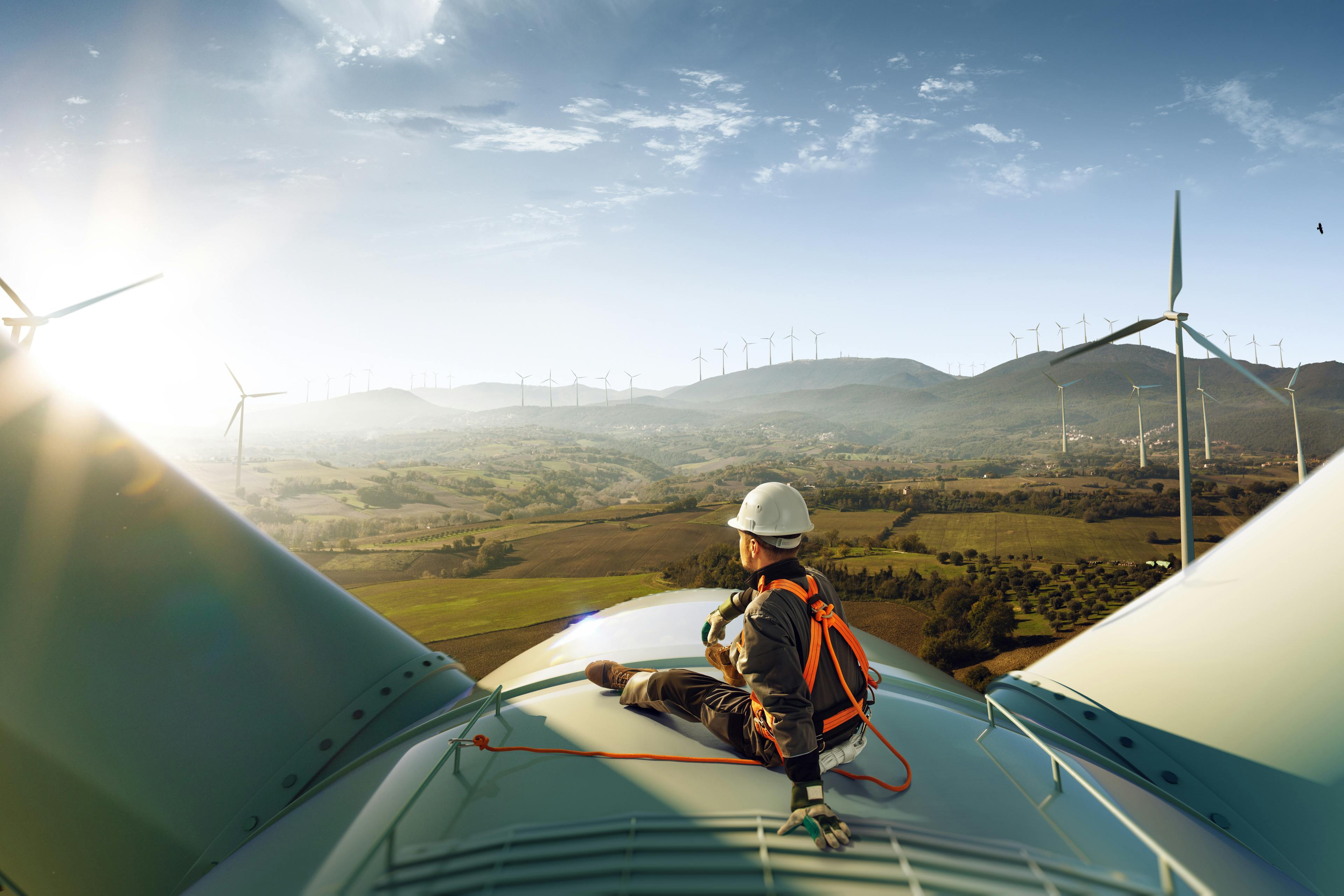 A man in a hard hat sits on top of a windmill in a windfarm.