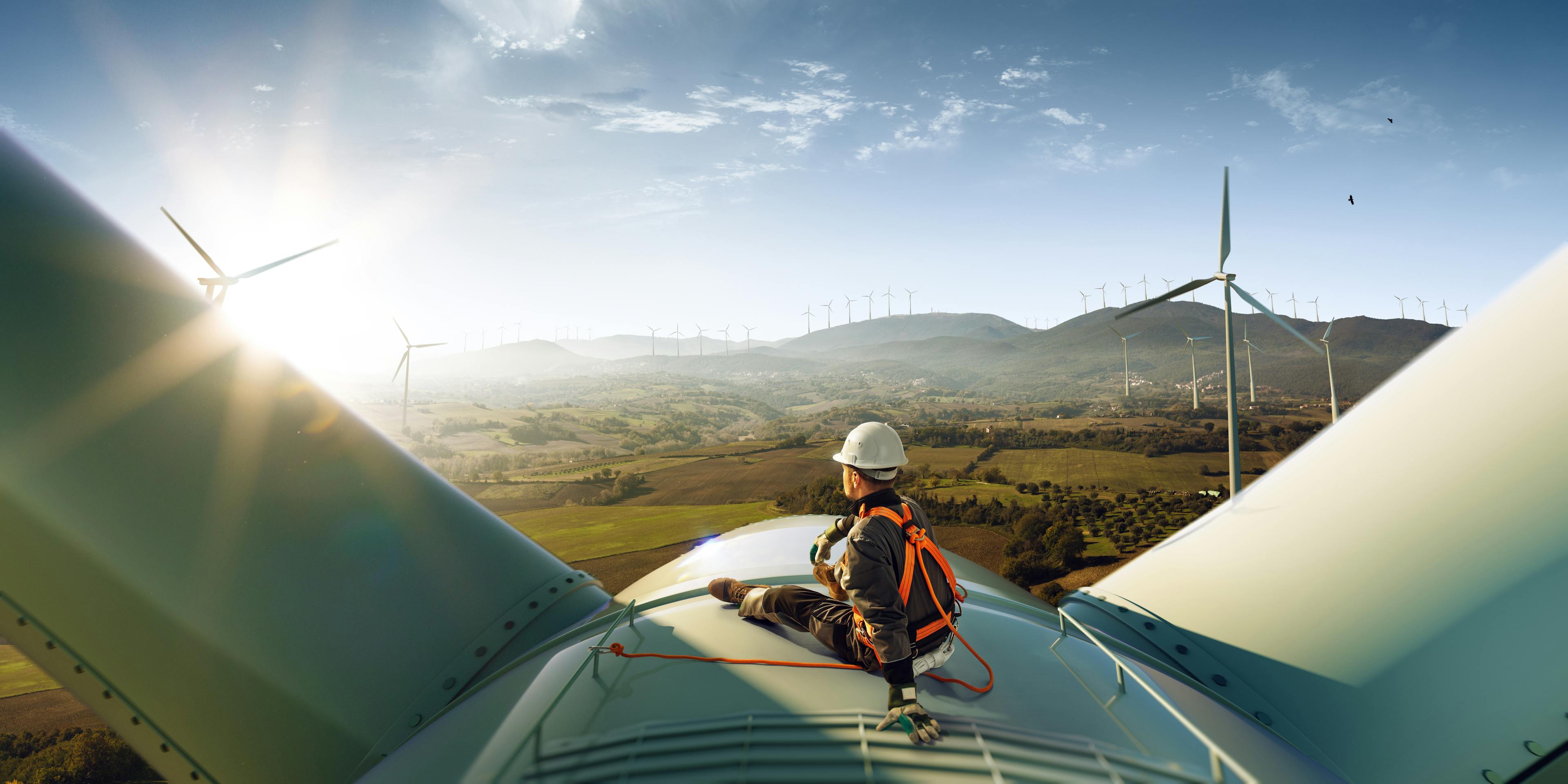 A man in a hard hat sits on top of a windmill in a windfarm.
