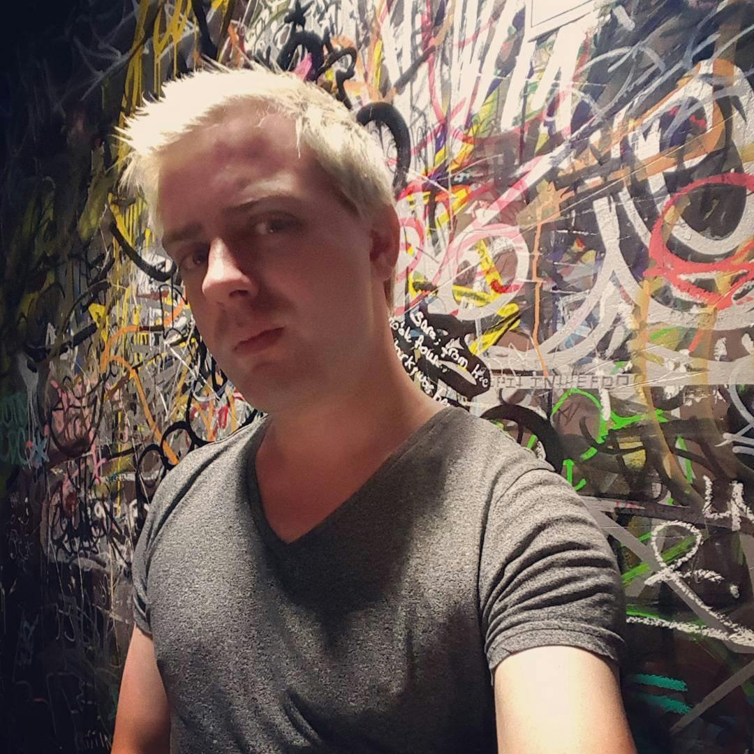 Medium shot photo of Alexis Watson, sporting their platinum blonde hair. A colorful wall of graffiti is in the background.