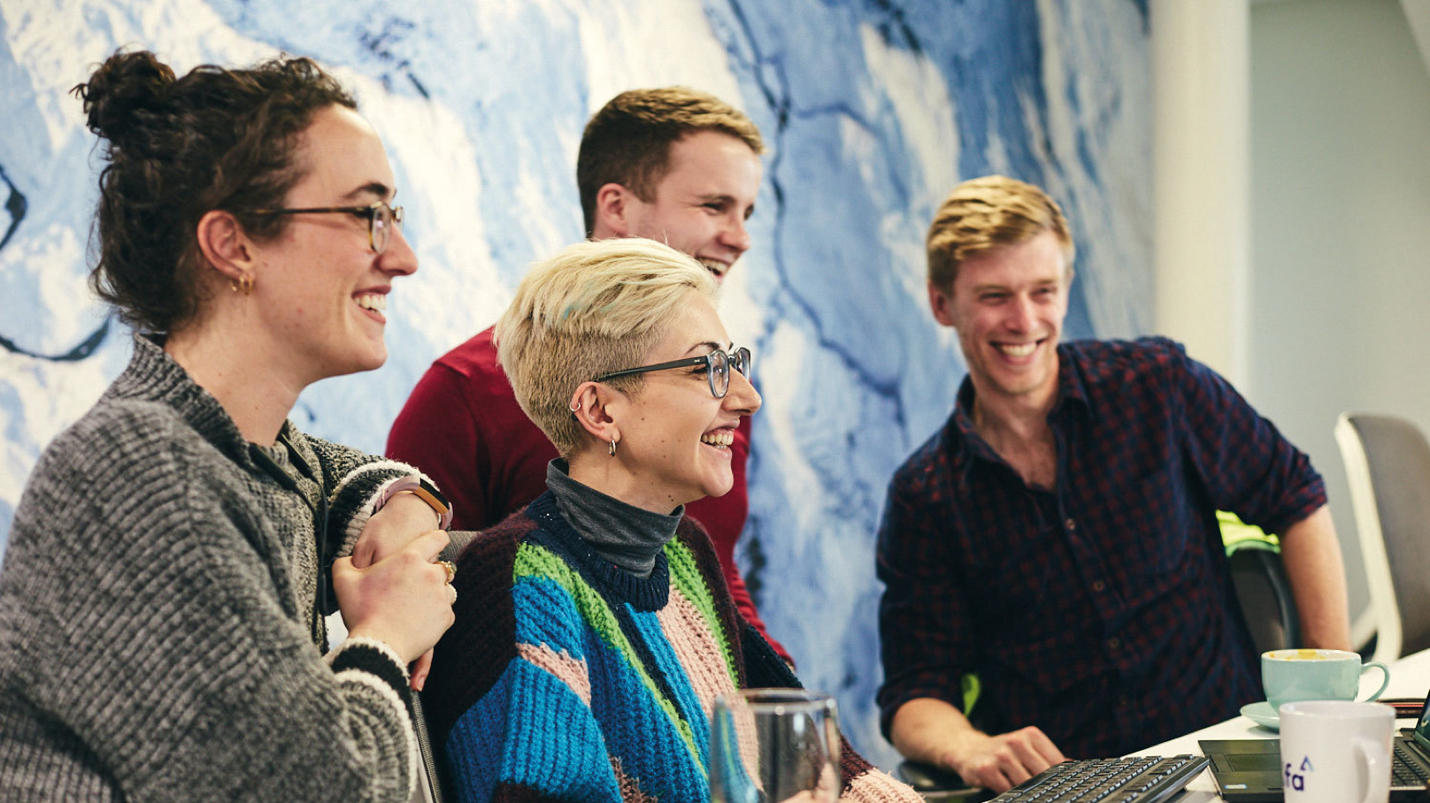 Four Alfa colleagues, two women and two men crowd and smile around a computer screen