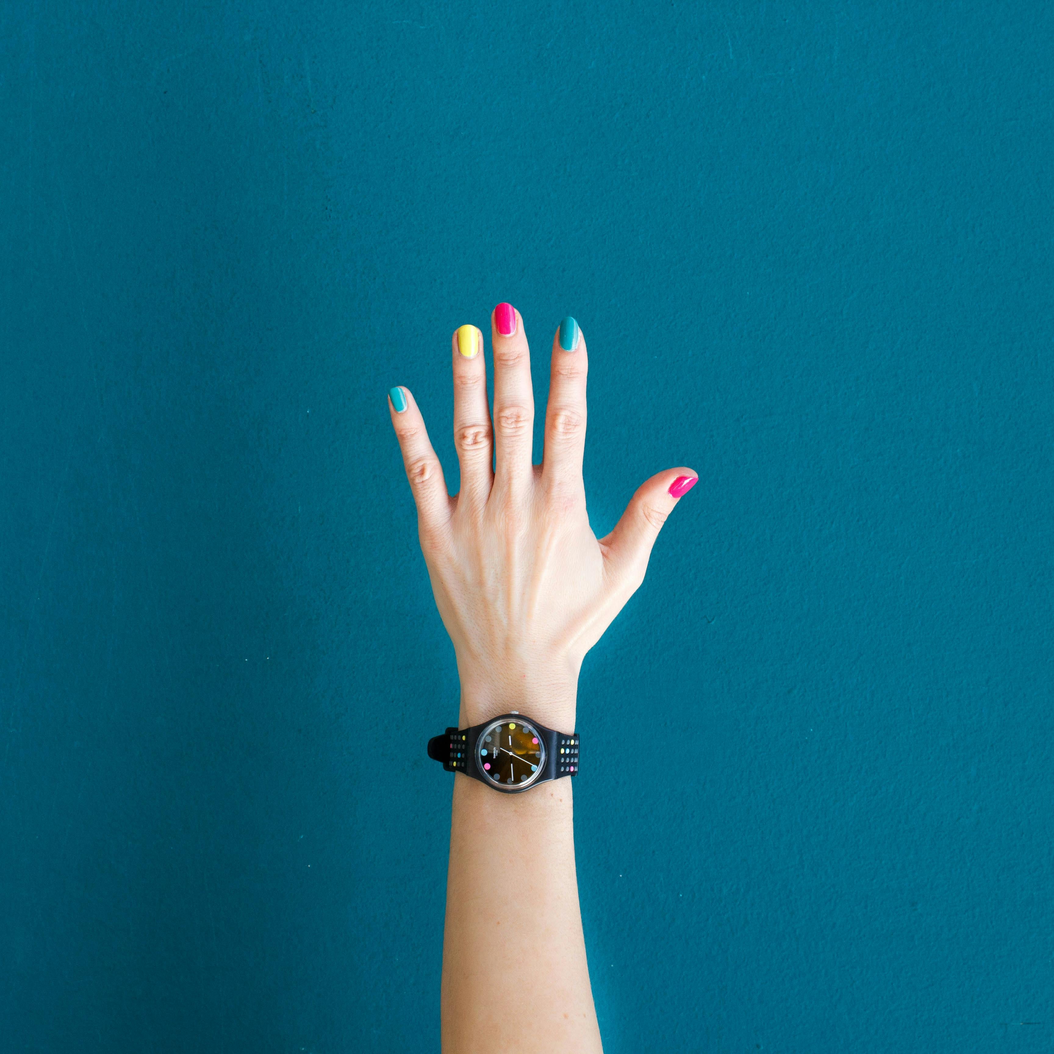 Raised hand with a black watch and multicolored nail polish on blue background