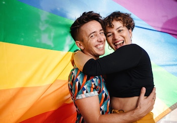 A smiling couple embraces in front of a Pride flag