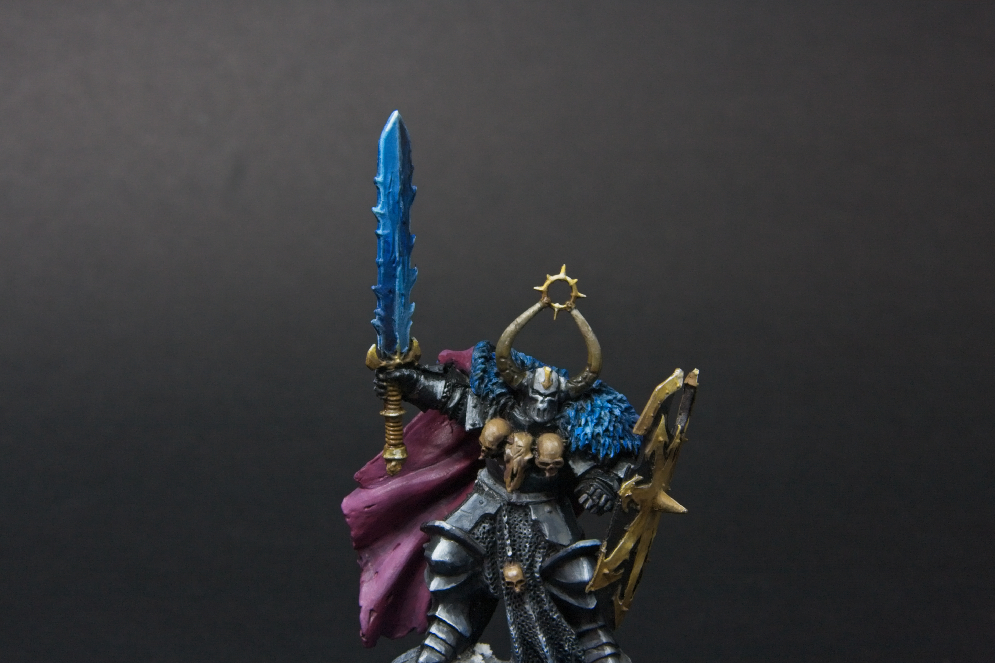 Old metal archon, with three skulls at his chest, a mauve cape, a blue sword, and a golden shield.