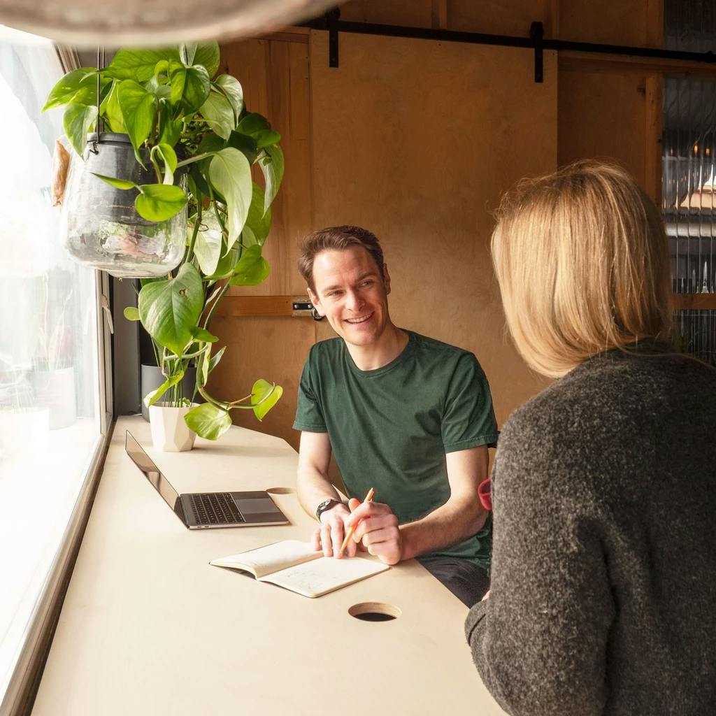 Image of Barney sat being interviewed by Katie (whose back is to the camera), writing in a notepad with his laptop open to the left of him.