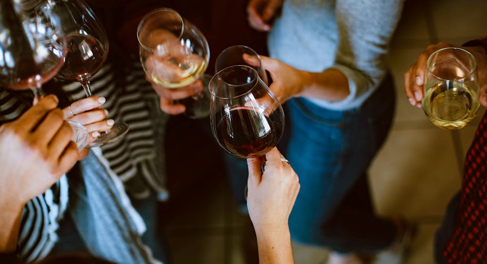 Image of hands holding wine glasses, taken from above - you can't see any faces. There's a mix of white and red wine in the six glasses you can see.