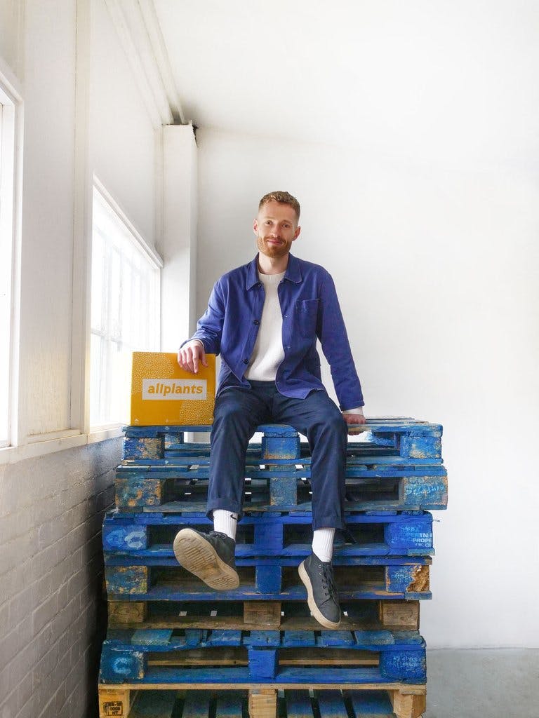 Image of our director of customer experience, Ferdie, sat on top of a stack of blue palettes wearing slack trousers, a white t-shirt, and an unbuttoned blue shirt. He's smiling with his right arm resting on top of an allplants delivery box.