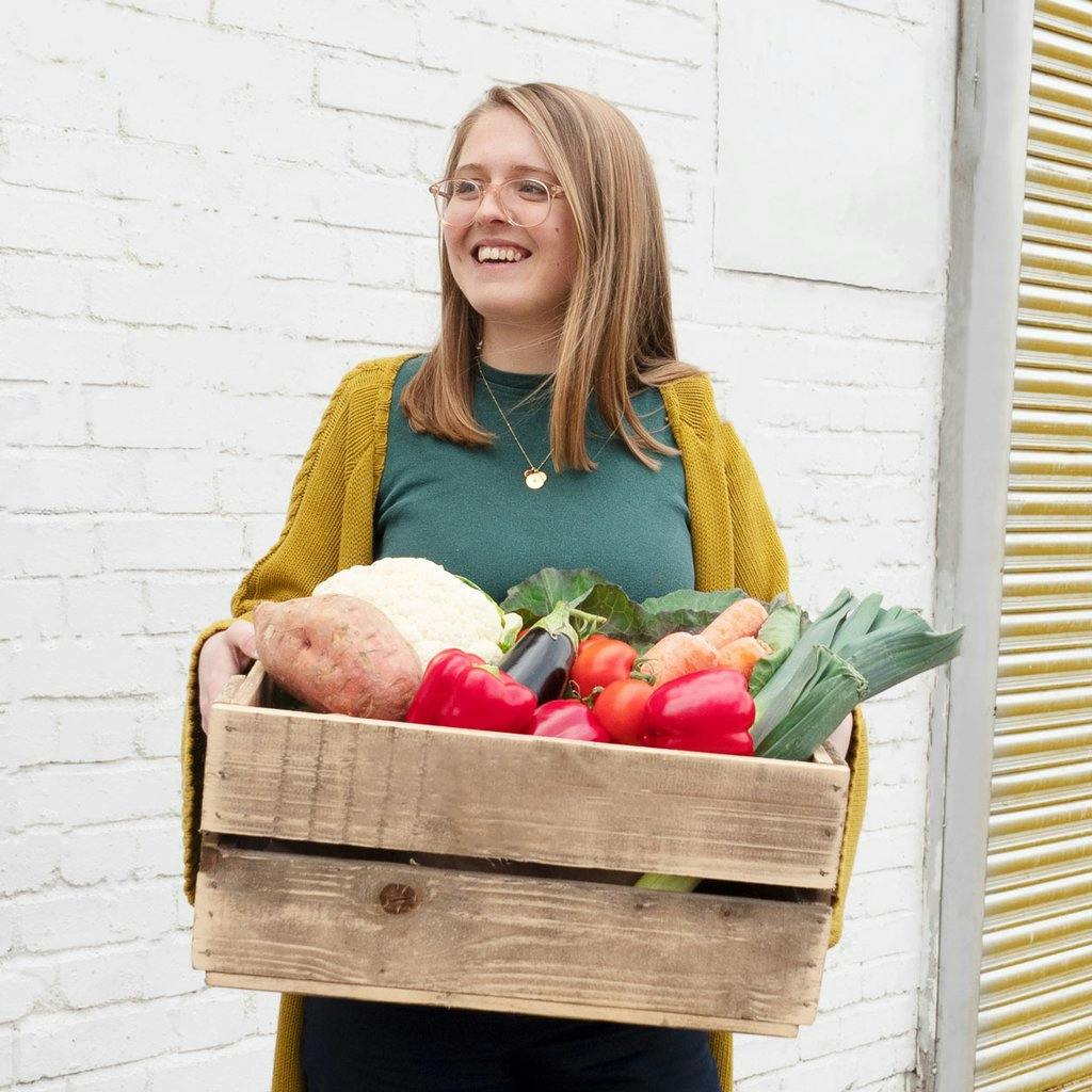 Image of Ellie holding a wooden crate full of red peppers, leeks, and cauliflower, stood in front of a white brick wall. She's wearing a green t-shirt and mustard cardigan, and is smiling and looking to the left away from the camera.