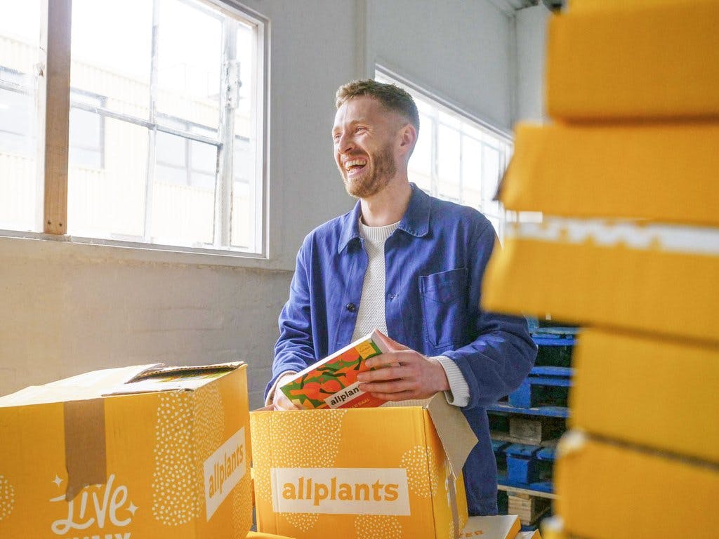 Image of ferdie behind a couple of stacks of allplants delivery boxes. He's wearing a white t-shirt with an unbuttoned blue shirt layered over the top. He's smiling, looking away from the camera to the left and is packing an allplants dish into an open allplants delivery box.