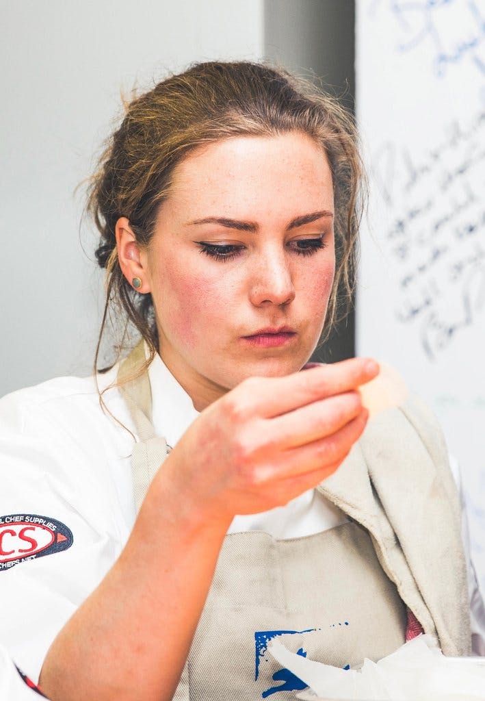 Close up imgae of Joey, our senior development chef, in her chefs whites and allplants apron. Shes looking at an ingredient that she's holding in her right hand hand which is raised up level with her jaw.