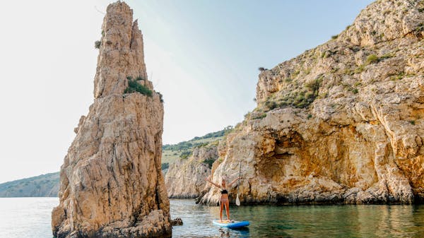 A paddleboarder stands on her board beside a stack of rocks in the waters off the rugged Krk island, Croatia