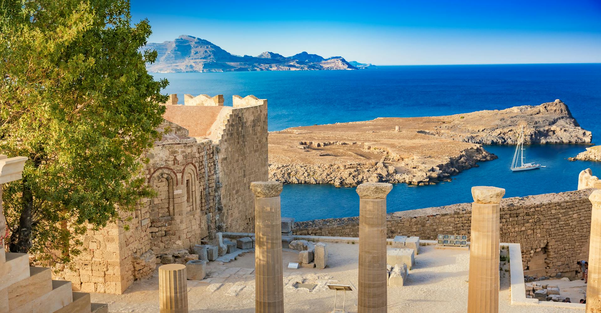 A yacht travels towards a Greek harbour, where stone pillars and ruins stand on a hill in the foreground