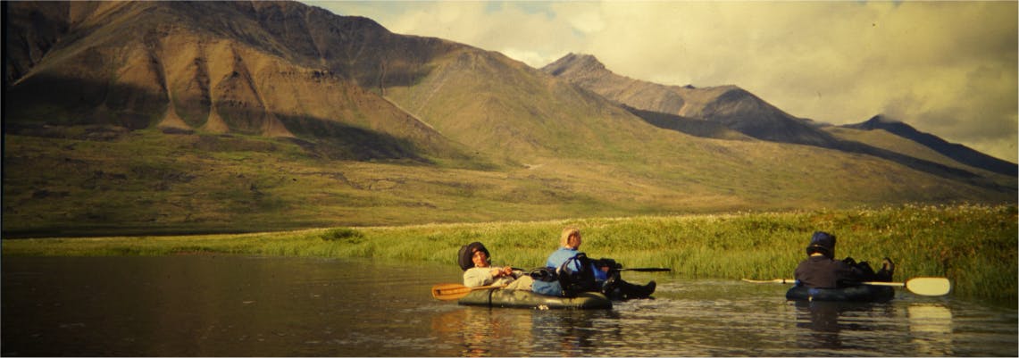 Scanned photograph of a group of people relaxing in water on packrafts with mountains in background