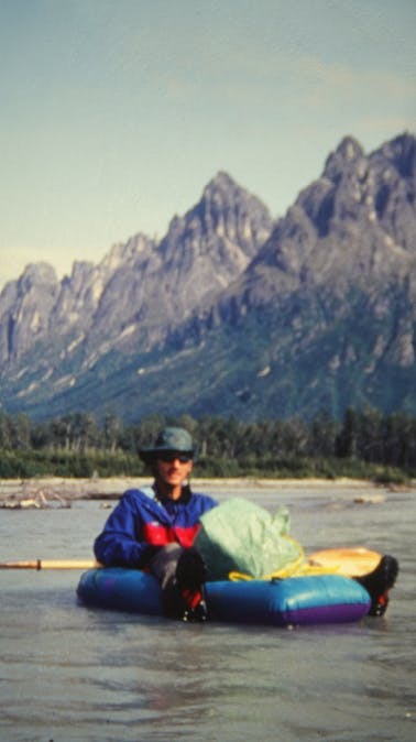 Scanned photograph of man in river sitting on packraft with large mountain range in background