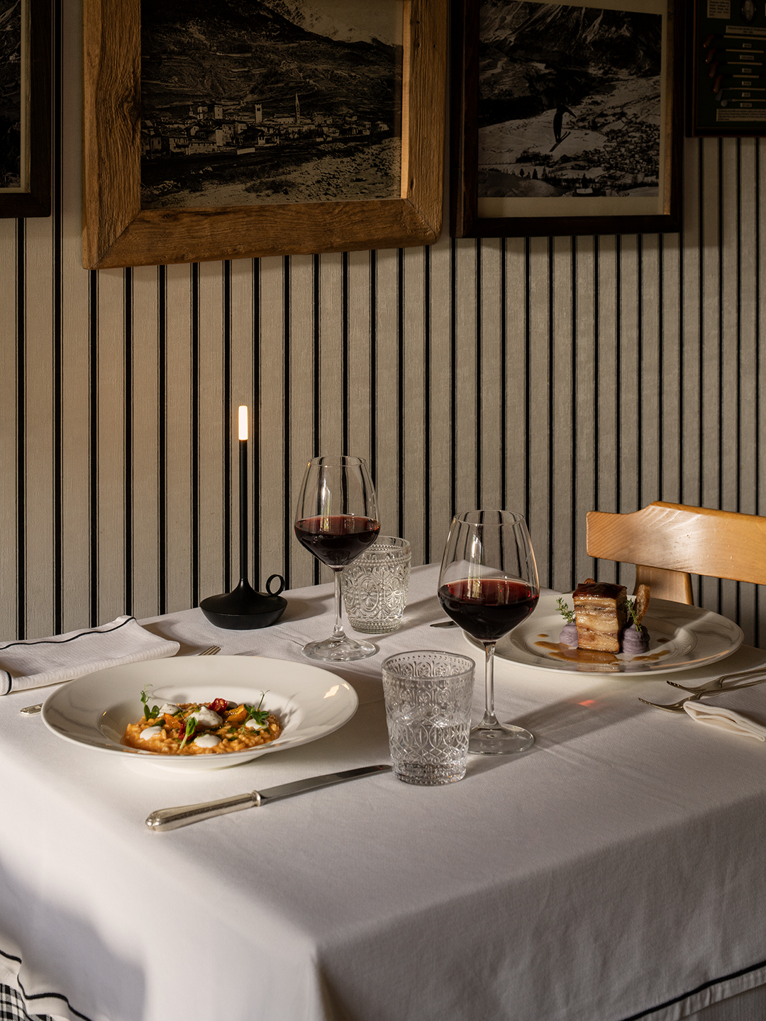 Tavolo ristorante con piatti gourmet, calici di vino rosso e candele accese, parete decorata con foto storiche d'epoca