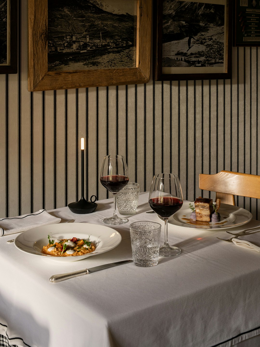 Tavolo ristorante con piatti gourmet, calici di vino rosso e candele accese, parete decorata con foto storiche d'epoca