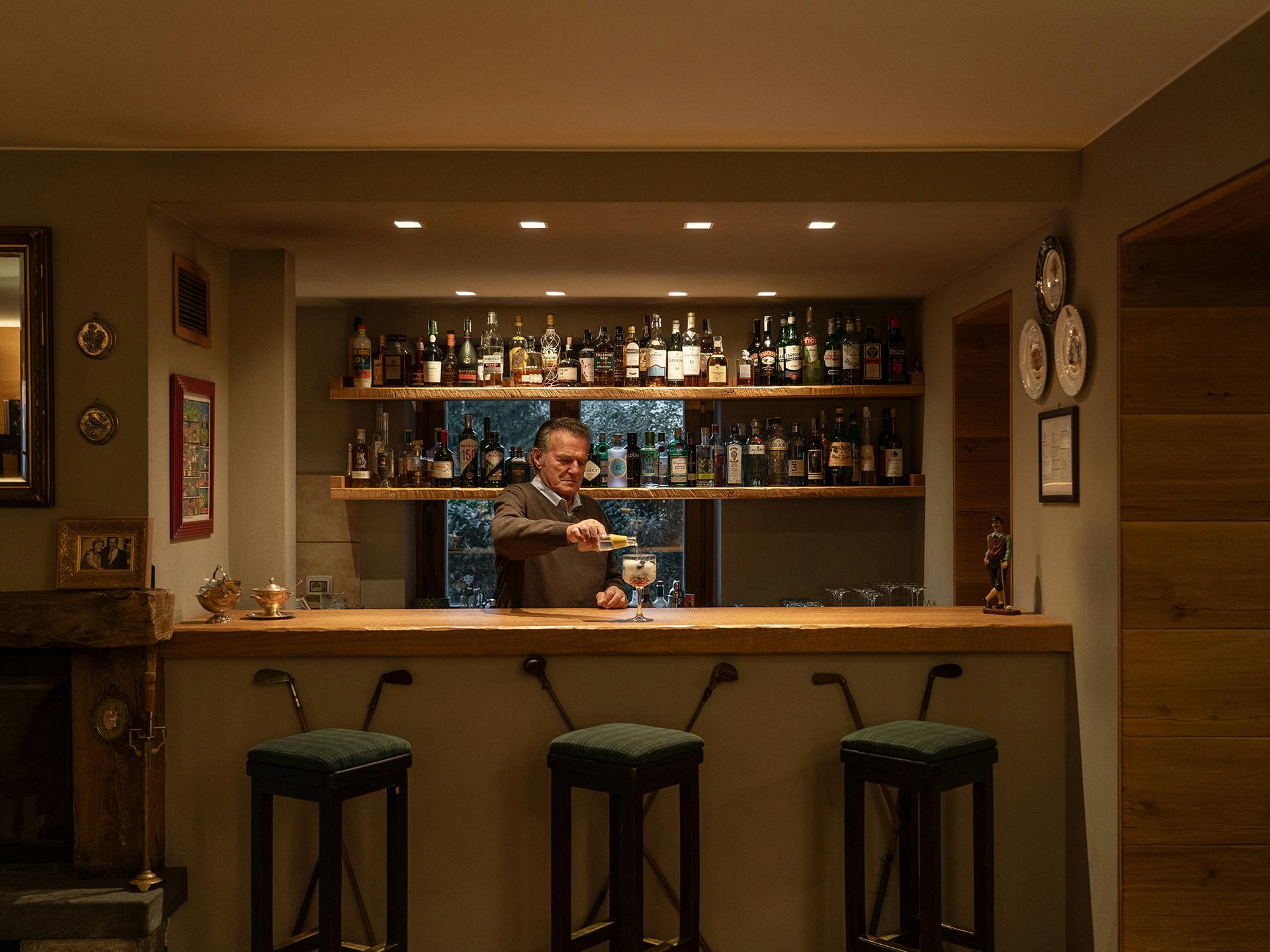 Proprietario al lavoro dietro bancone bar elegante con ricca selezione di liquori su mensole illuminate, sgabelli rivestiti in velluto verde