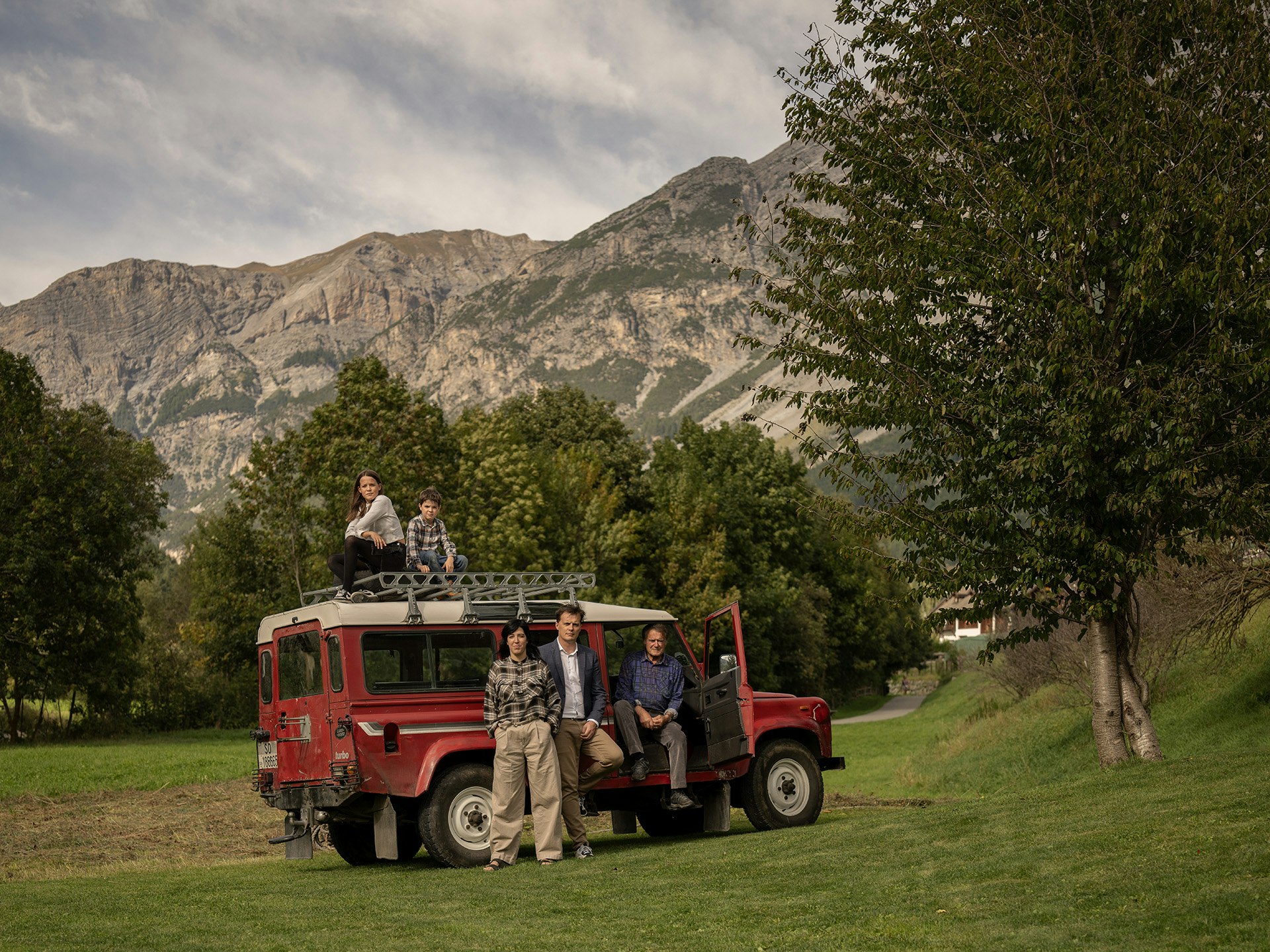 Foto di famiglia con Land Rover d'epoca rosso su prato alpino con vista sulle montagne