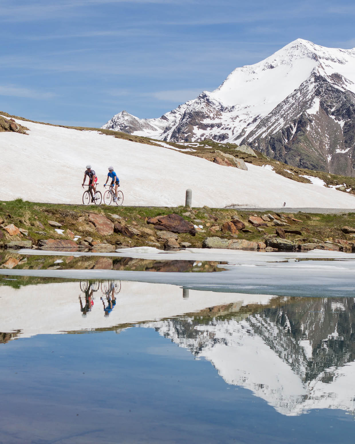 Ciclisti in alta montagna riflessi in pozza d'acqua, paesaggio alpino con neve e cime innevate