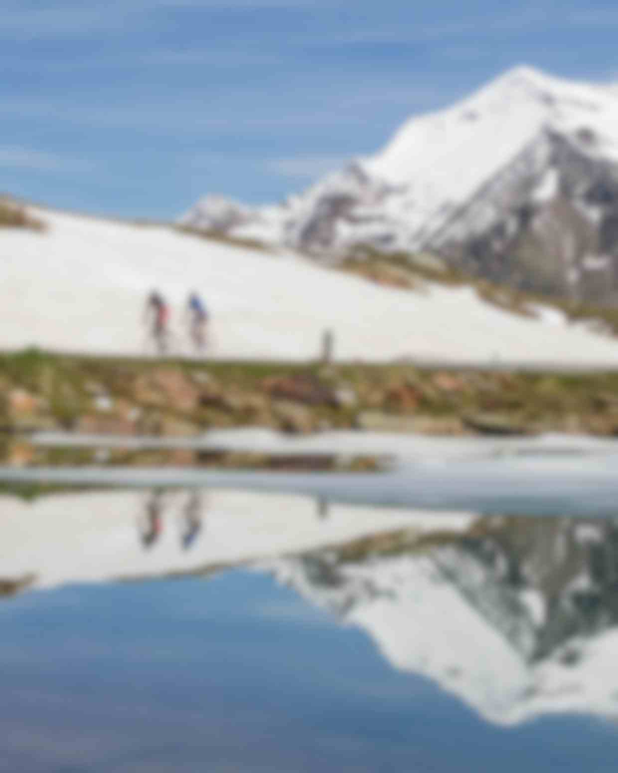 Ciclisti in alta montagna riflessi in pozza d'acqua, paesaggio alpino con neve e cime innevate