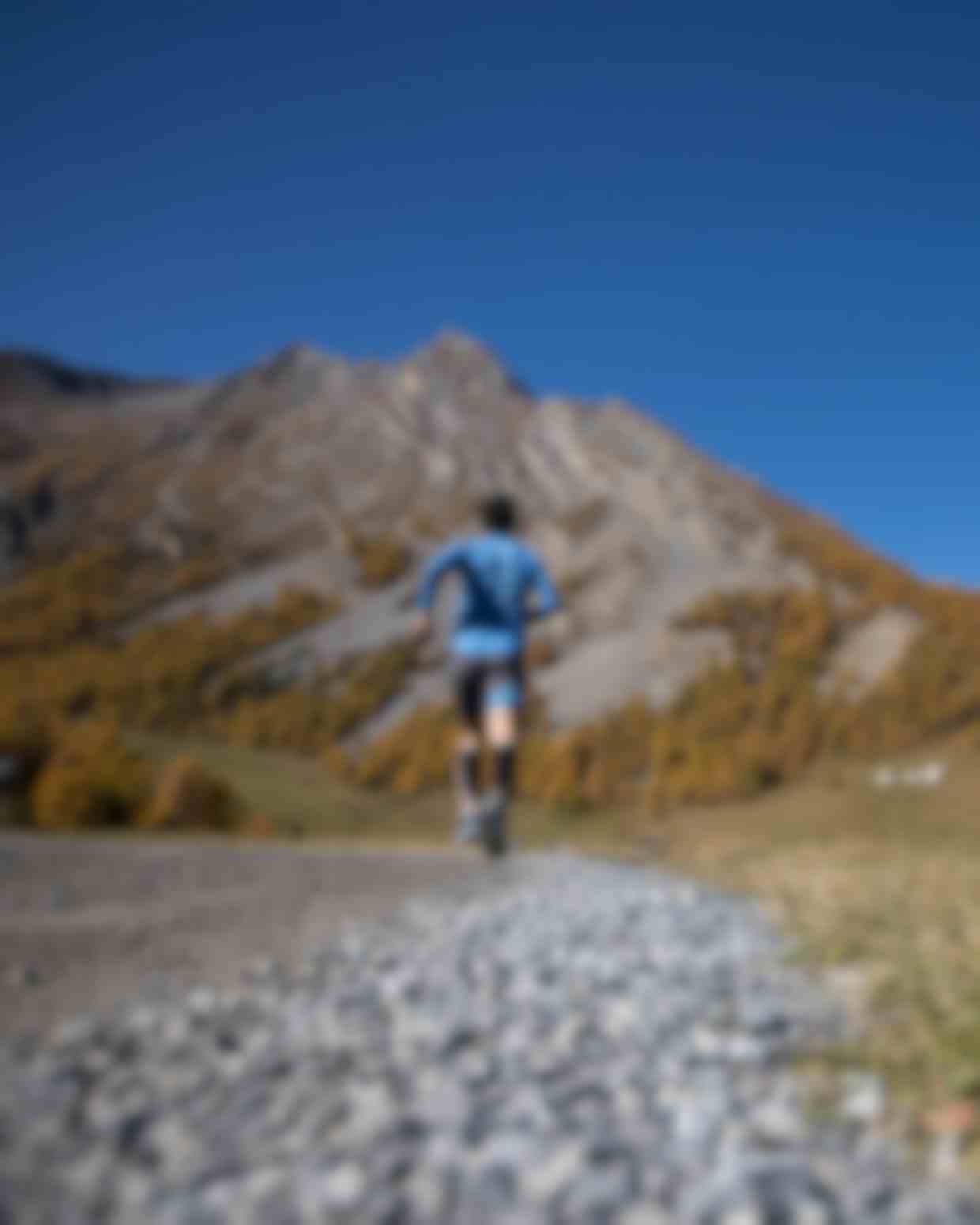 Trail runner su sentiero di montagna con larici autunnali dorati e cime alpine sullo sfondo