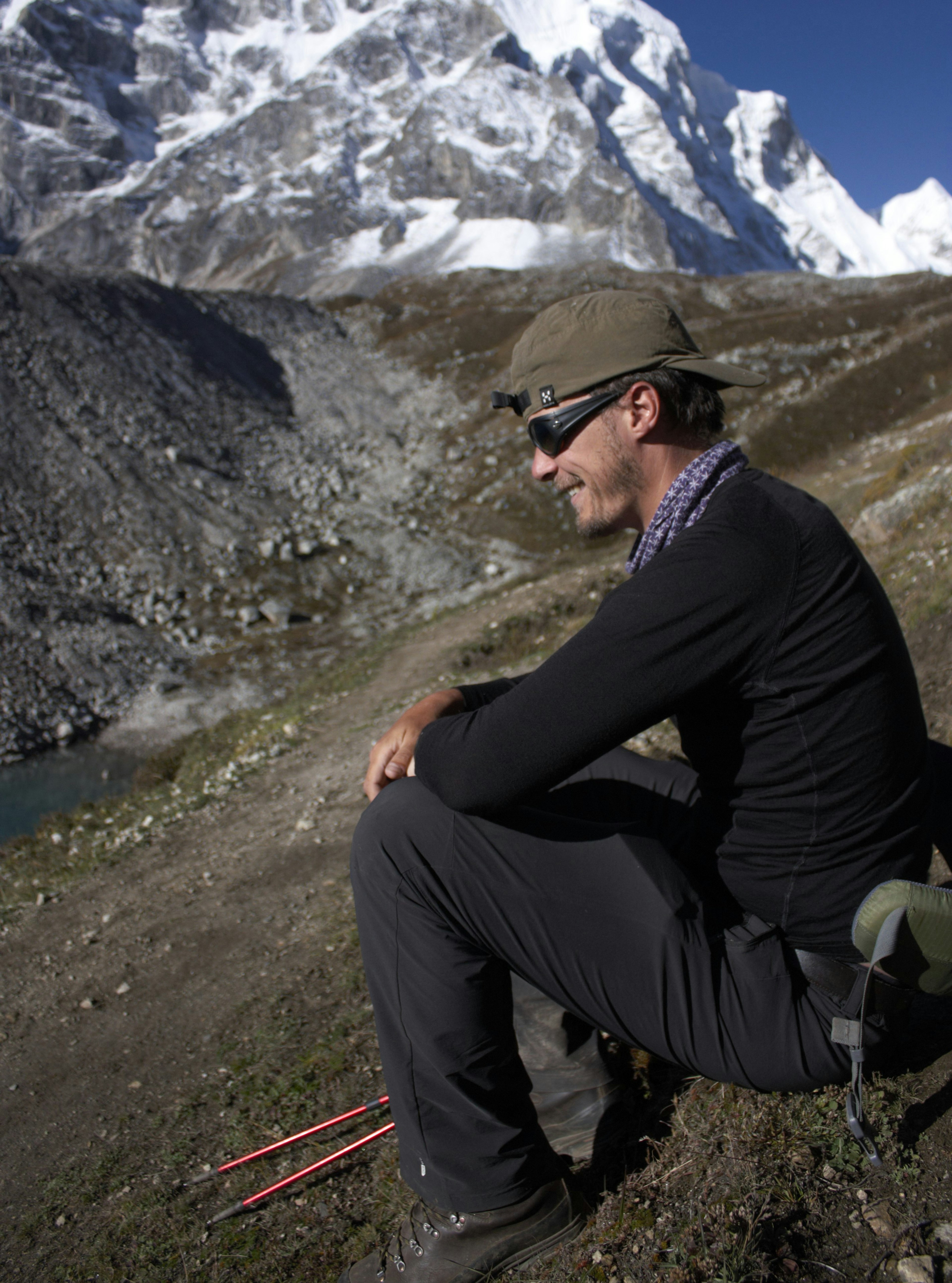 Escursionista seduto su roccia contemplando lago alpino e cime innevate, bastoncini da trekking in primo piano