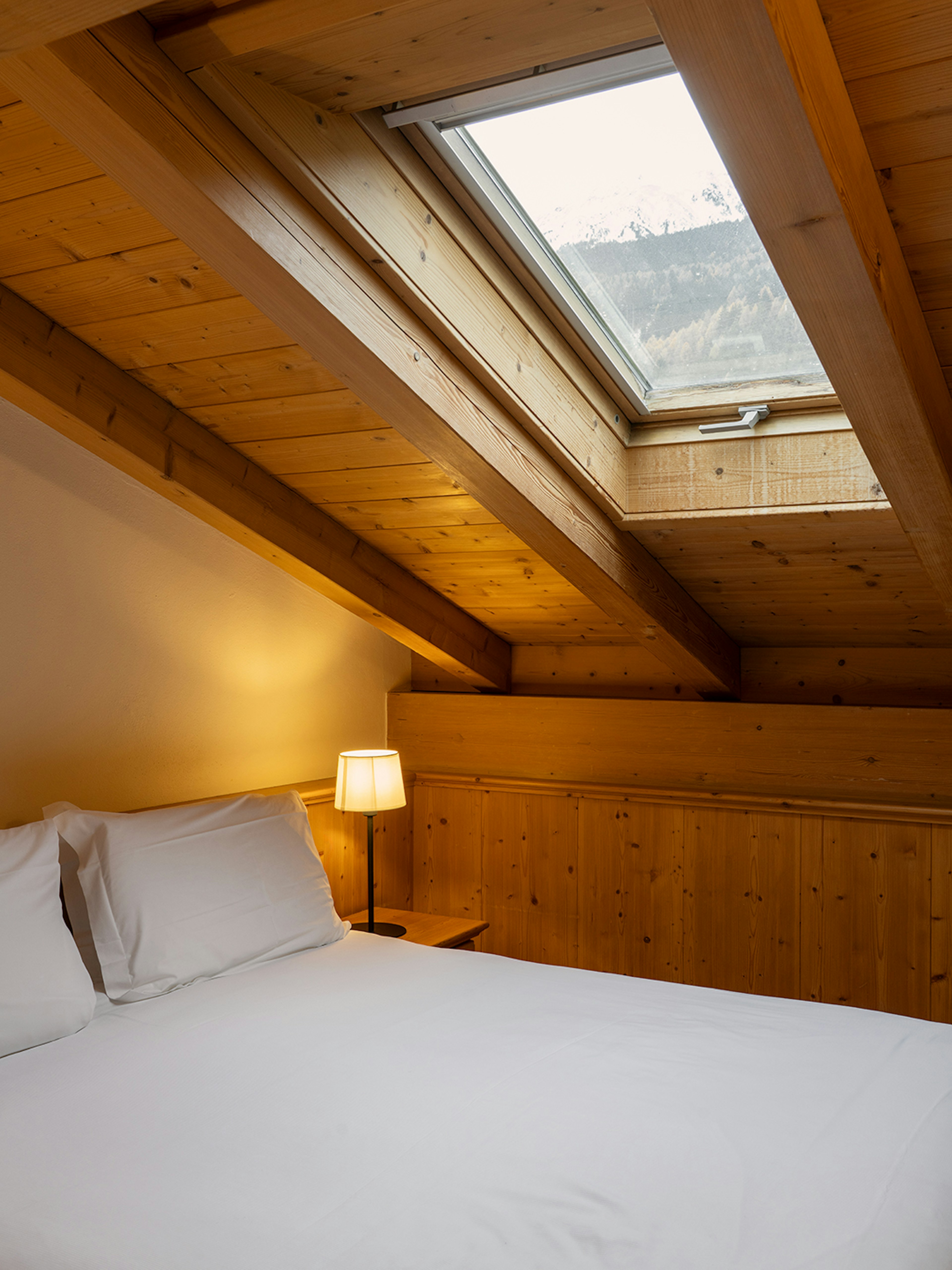 Camera mansardata in legno chiaro con letto singolo sotto il tetto spiovente. Una finestra sul tetto lascia entrare la luce naturale, mentre una lampada da comodino crea un’atmosfera calda e raccolta.