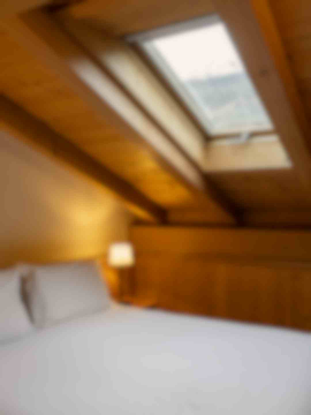 Camera mansardata in legno chiaro con letto singolo sotto il tetto spiovente. Una finestra sul tetto lascia entrare la luce naturale, mentre una lampada da comodino crea un’atmosfera calda e raccolta.