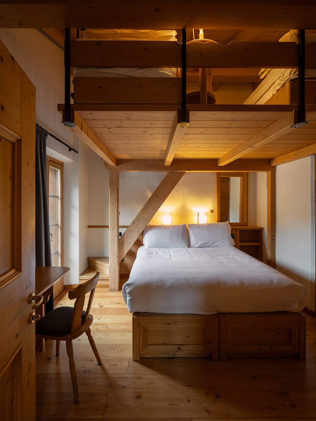 Camera in stile alpino con letto matrimoniale incorniciato da travi in legno a vista. Pavimento e arredi in legno chiaro, scrivania vicino alla finestra e illuminazione soffusa che rende l’ambiente accogliente